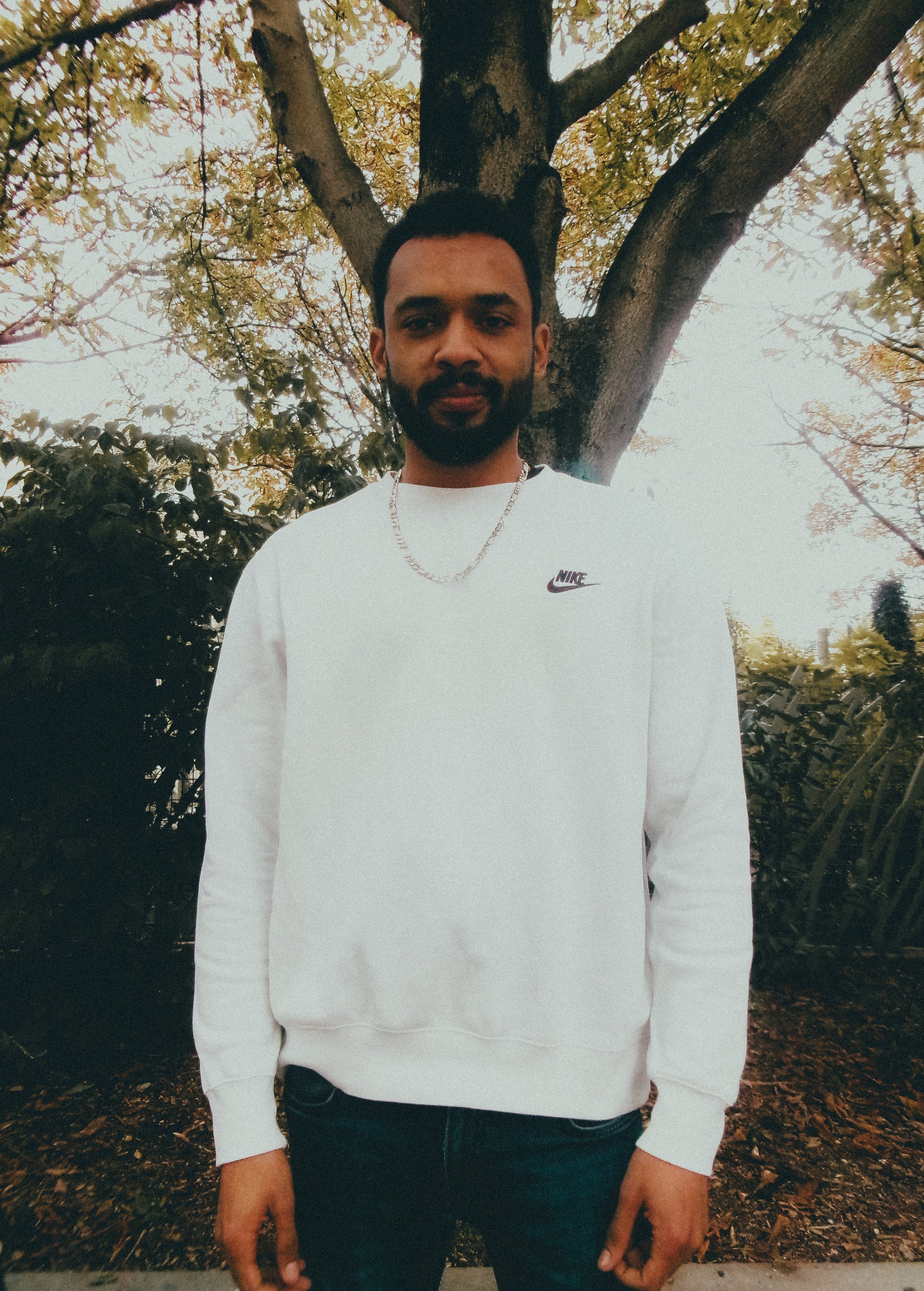 Jeune homme avec barbe, portant un sweat blanc Nike et une chaîne en métal, debout devant un arbre aux feuilles automnales dans un parc.