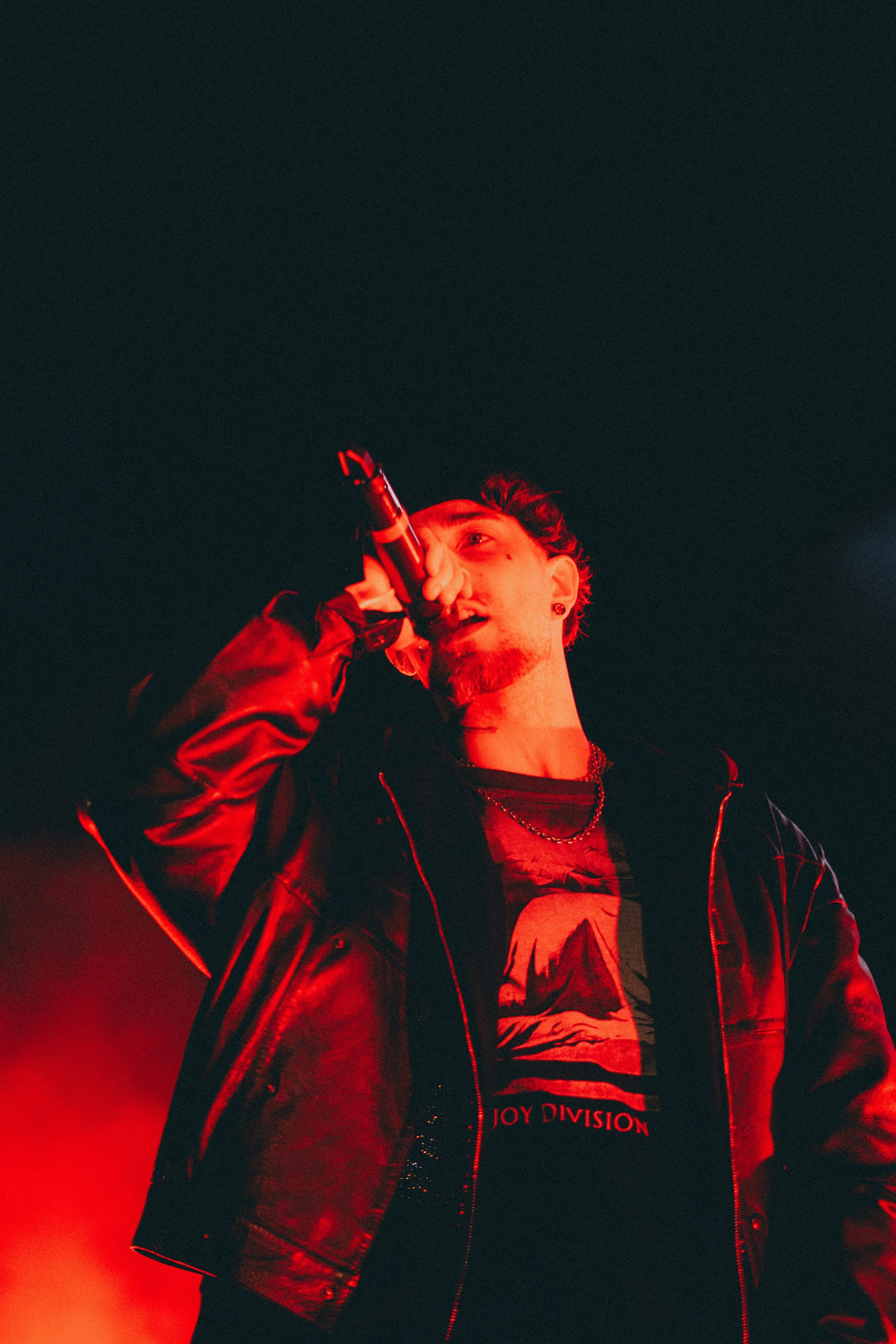Un homme chante dans un micro, portant une veste en cuir noir, éclairé par une lumière rouge, portant un t-shirt Joy Division.