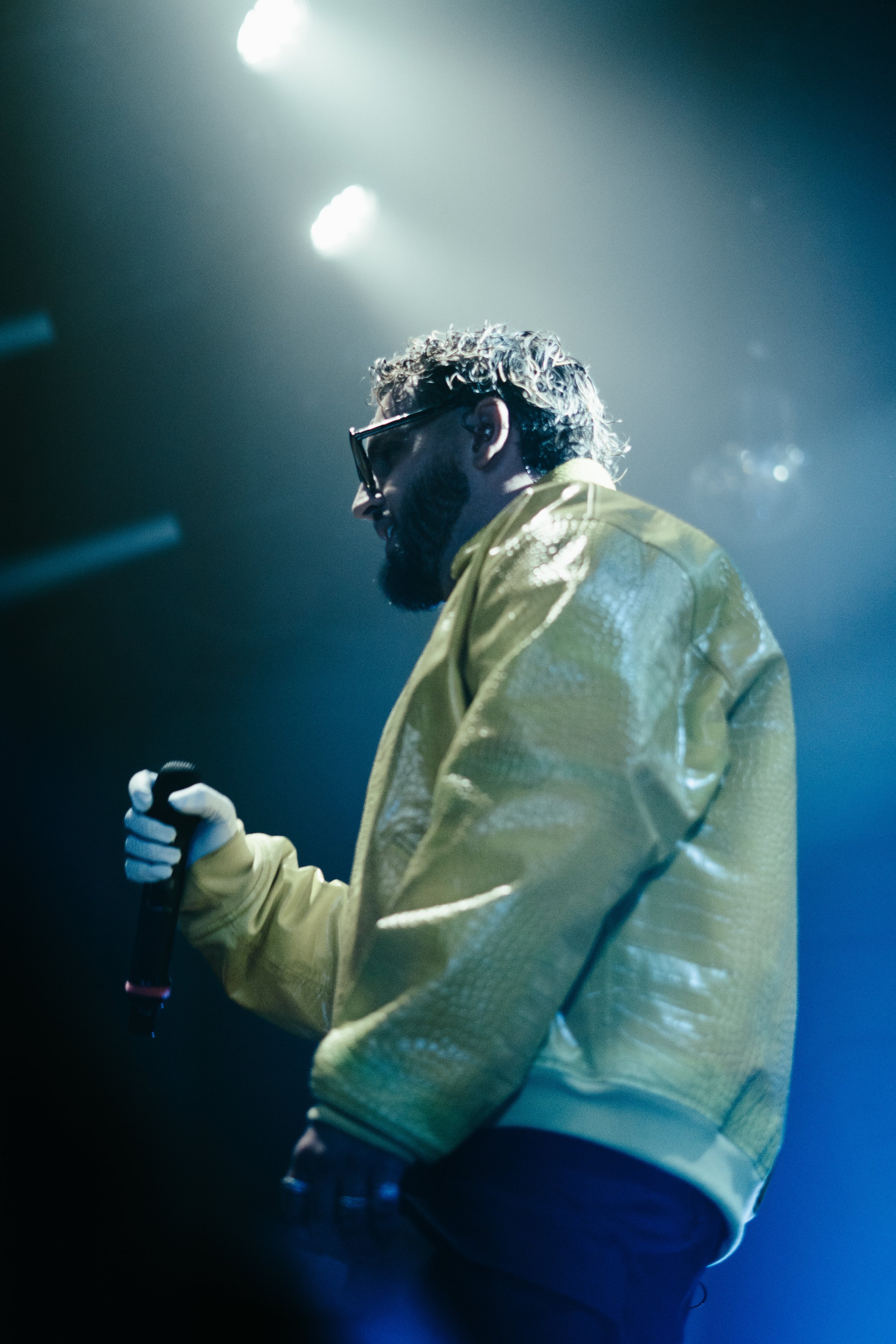 Un homme portant une veste jaune et des lunettes de soleil, tenant un microphone, sur une scène avec un éclairage bleu et blanc.