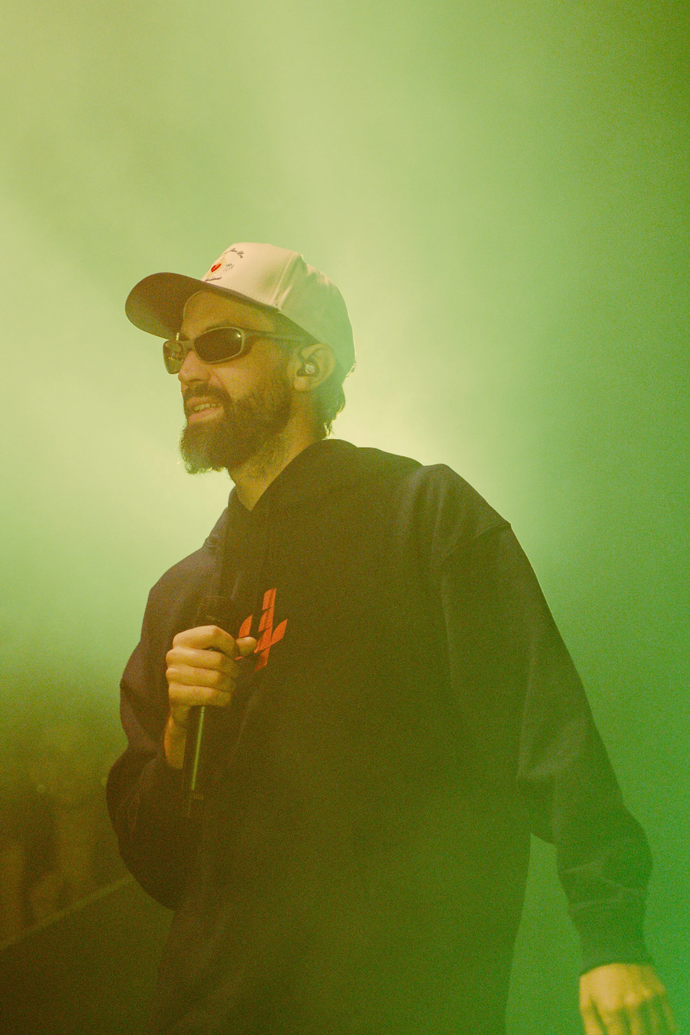 Un homme avec une barbe portant une casquette, des lunettes de soleil, un écouteur sans fil, et une veste noire, tenant un microphone, en scène avec un éclairage vert.