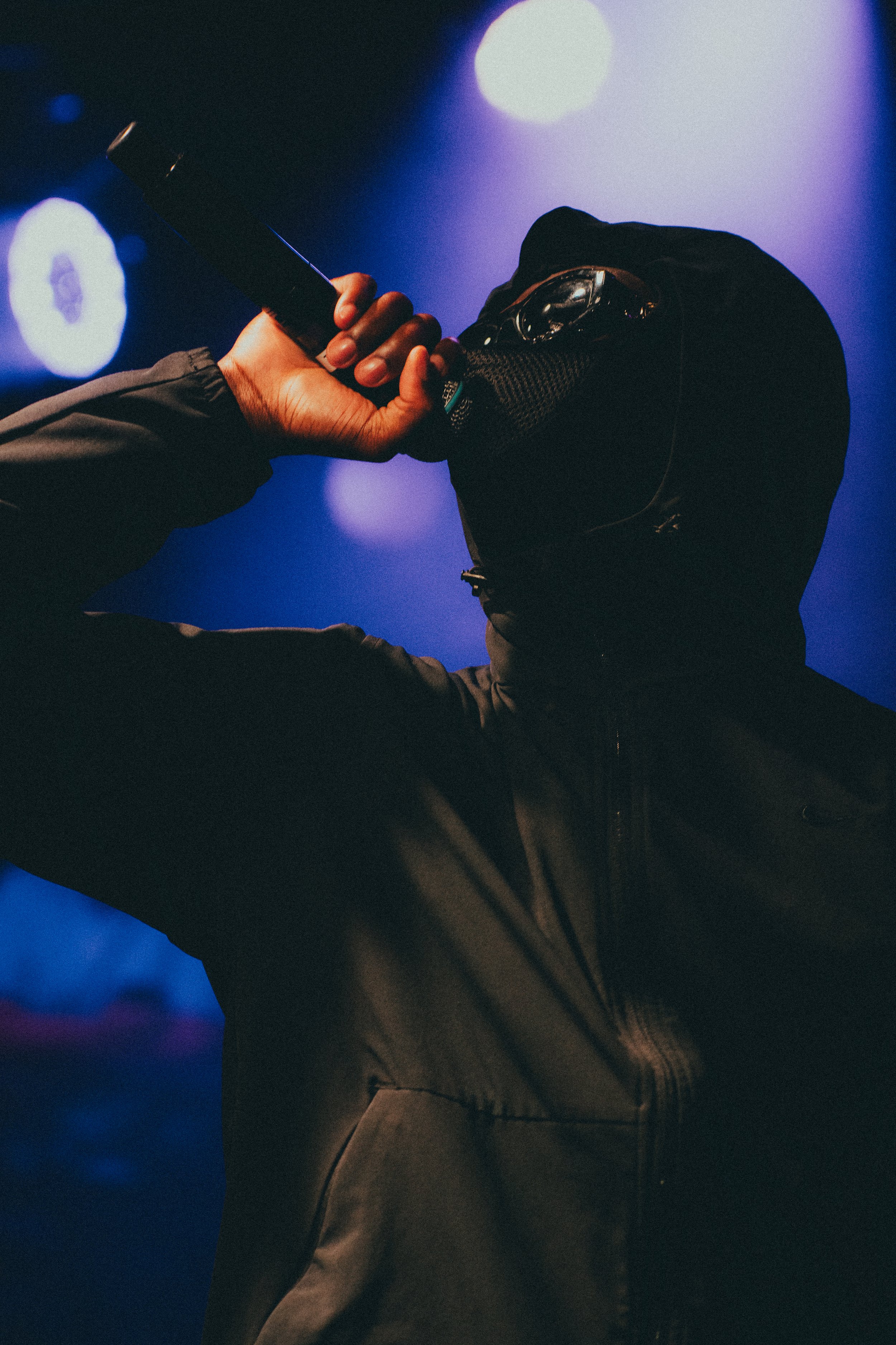 Un homme portant une capuche noire et un masque noir tient un couteau à la main, dans un environnement sombre avec un éclairage violet et bleu.