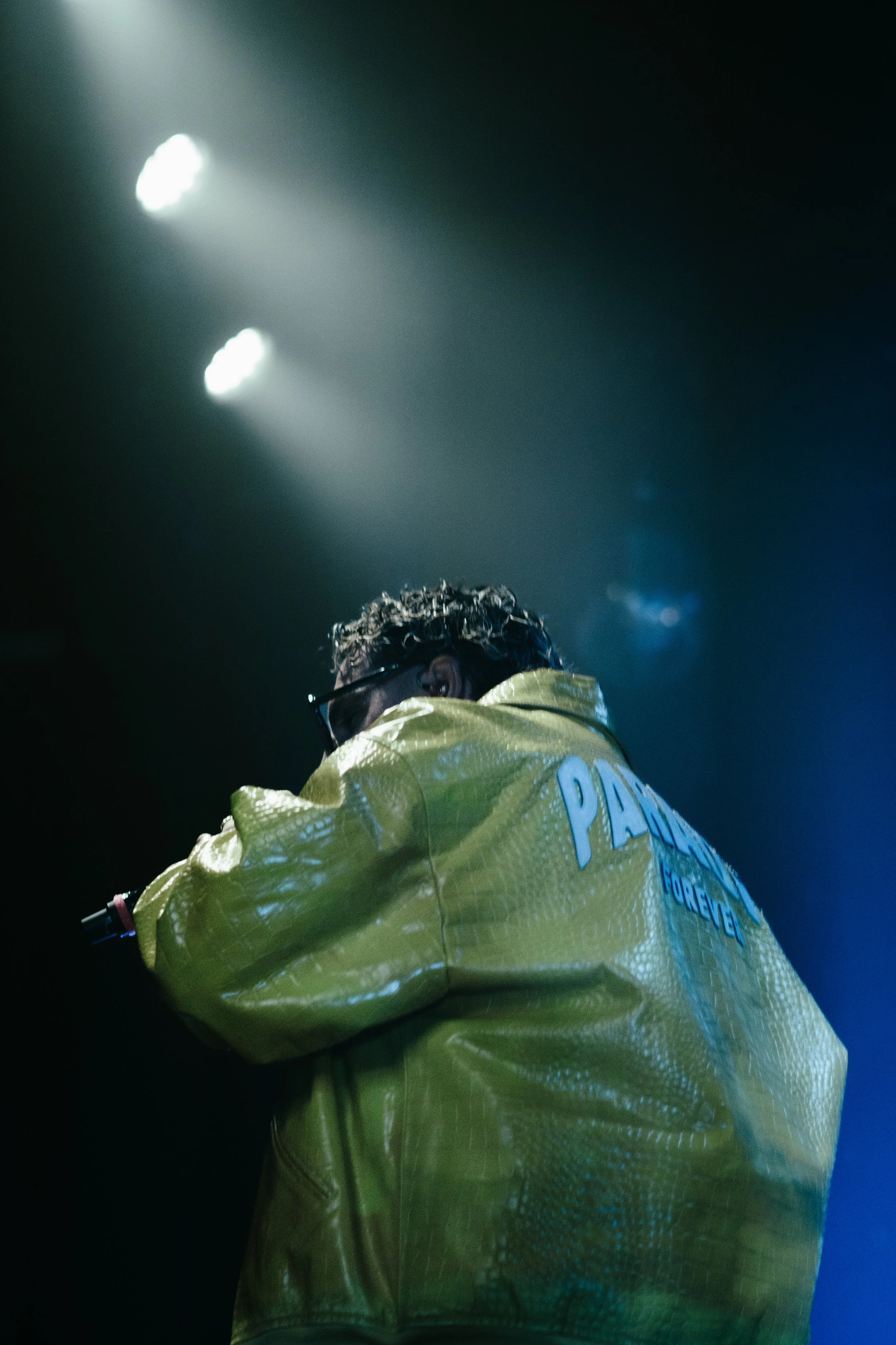 Personne portant une veste jaune avec le mot "PARADISE" écrit dessus, vue de dos dans un environnement sombre avec des lumières en arrière-plan.