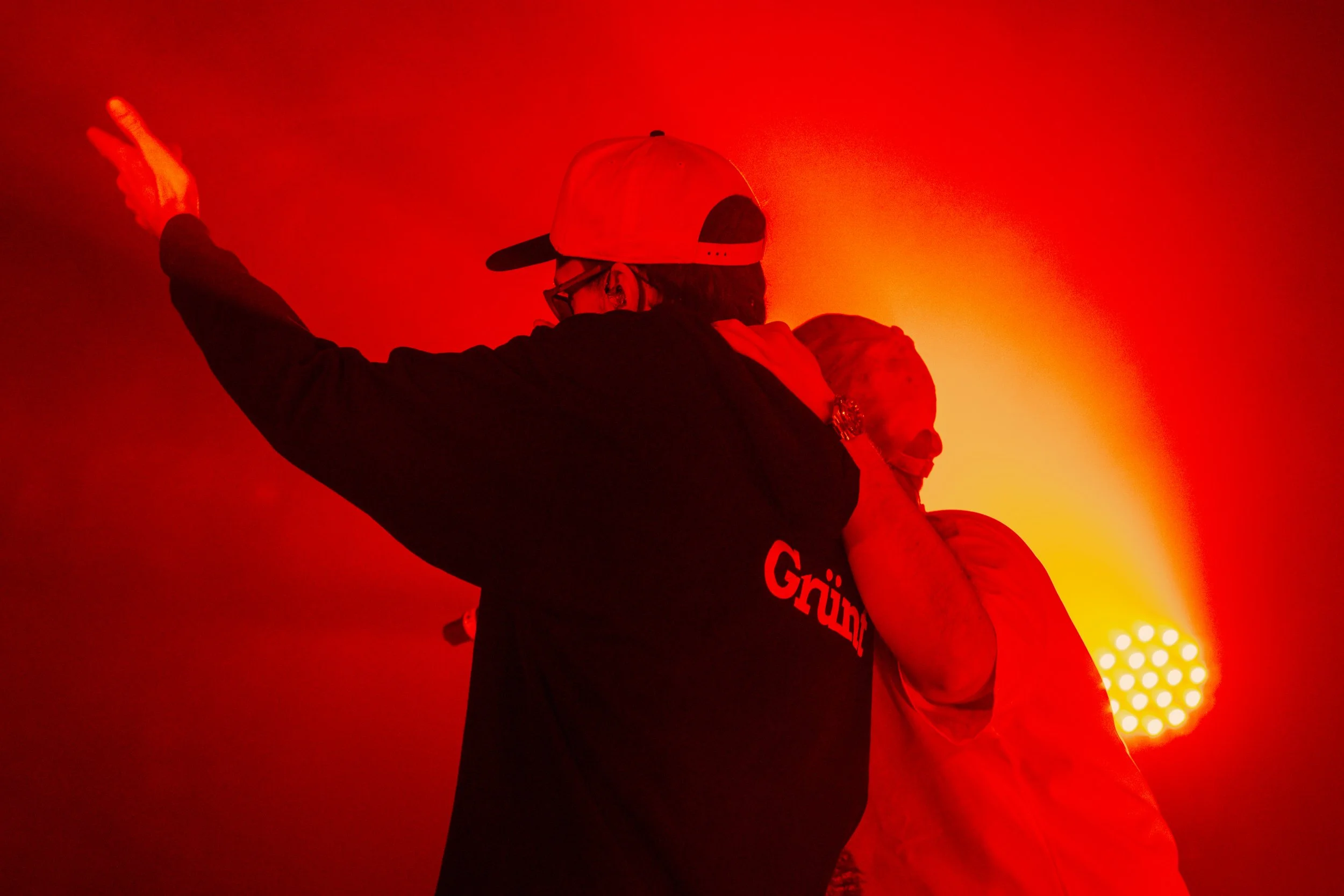 Deux personnes sur scène sous un éclairage rouge, l'une portant une casquette et un sweat avec le mot 'Gruum'.