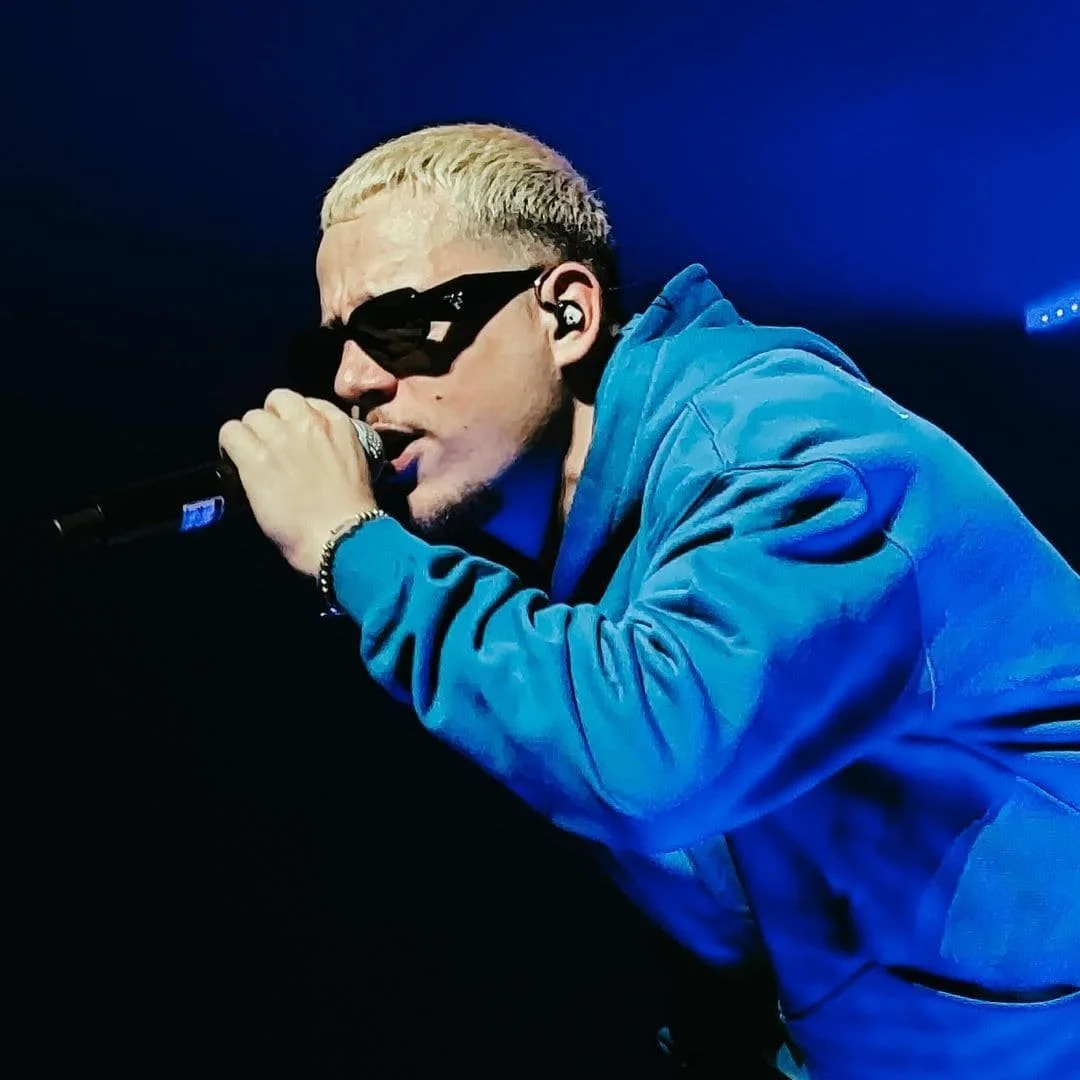 PLK portant des lunettes de soleil noires chante dans un microphone lors d'une performance, vêtu d'une veste bleue, sous un éclairage sombre avec une lumière bleue en arrière-plan.