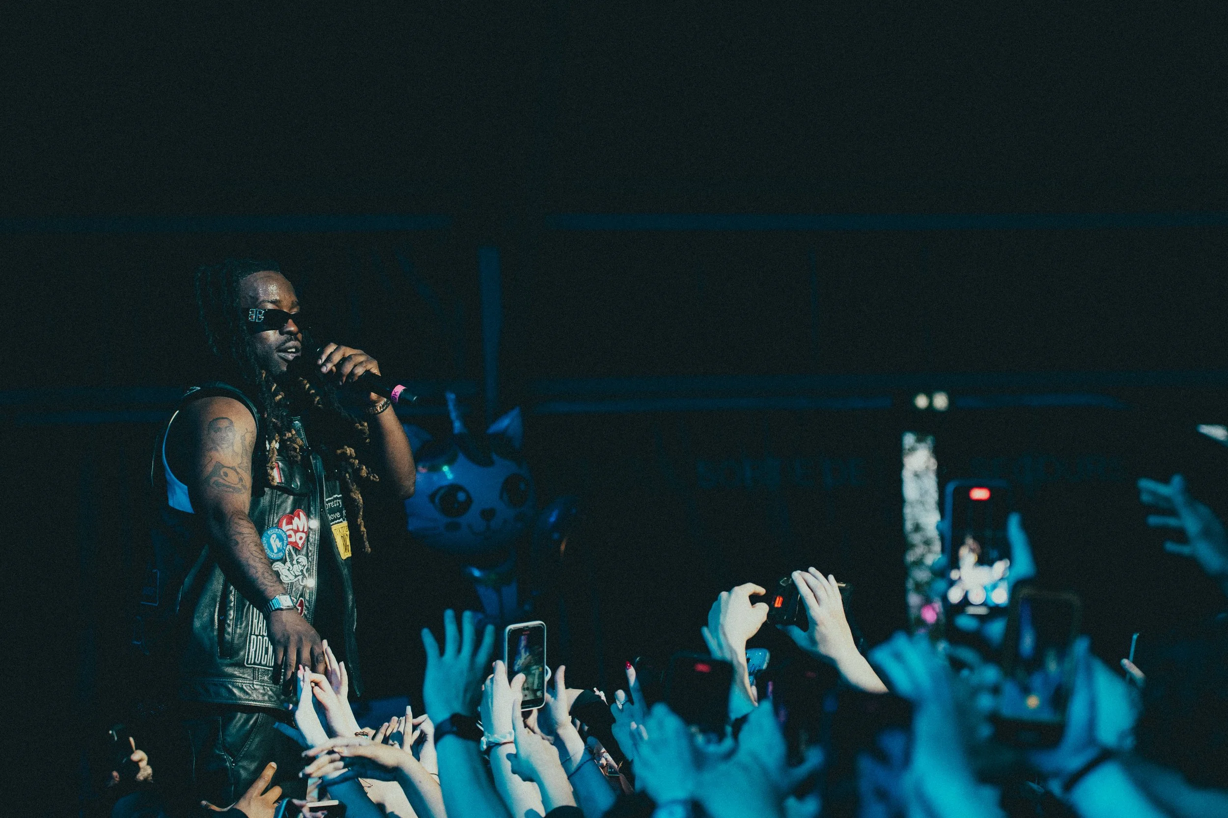 Un artiste de hip-hop en veste à patchs, portant des lunettes de soleil, tenant un microphone sur scène lors d'un concert. Audience avec mains levées et plusieurs personnes prenant des photos ou vidéos avec leur téléphone.