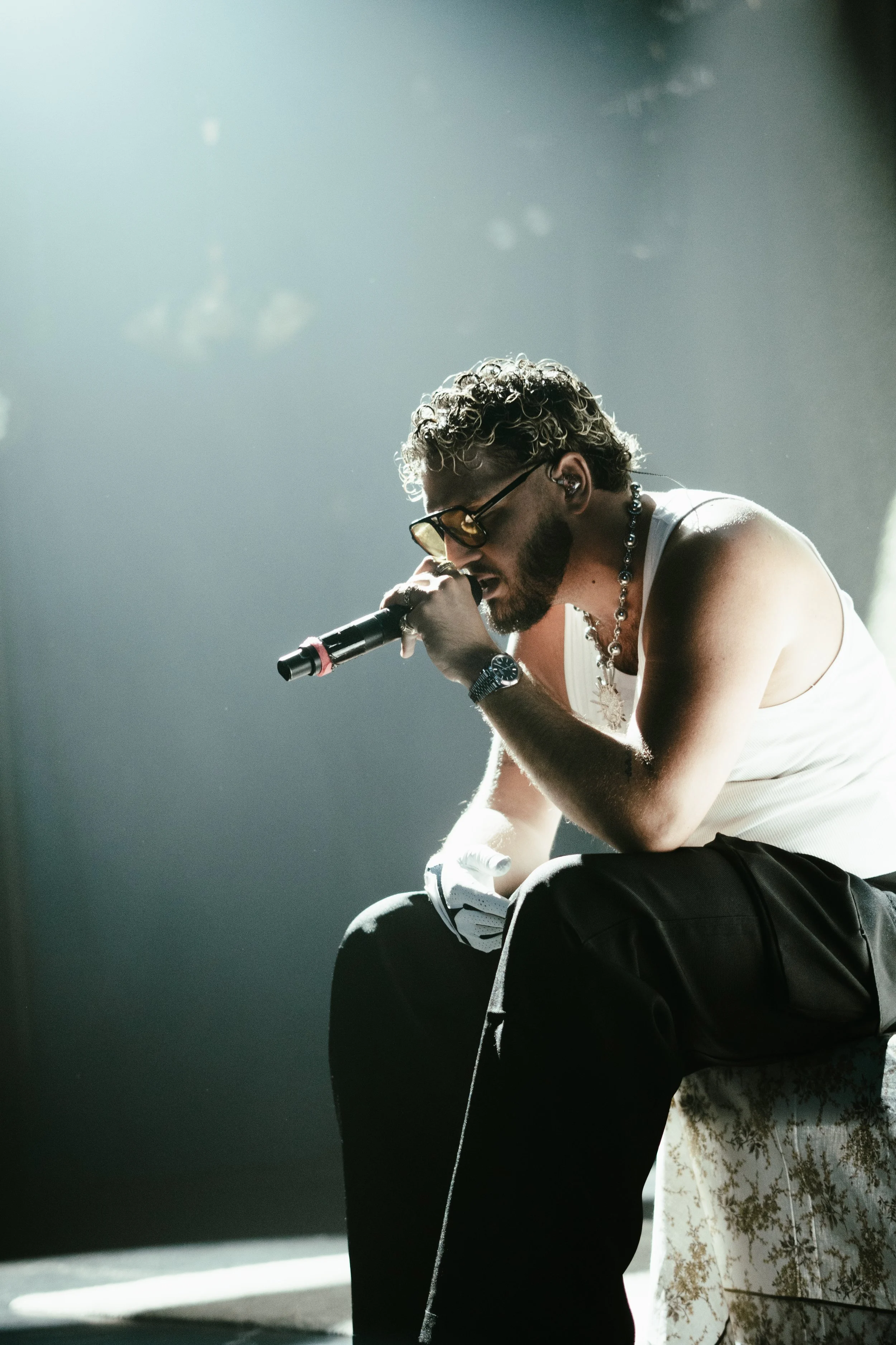 Un homme avec des cheveux bouclés, portant des lunettes de soleil, un collier, un bracelet, une montre, un t-shirt blanc et un pantalon noir, en train de chanter avec un micro sur scène.