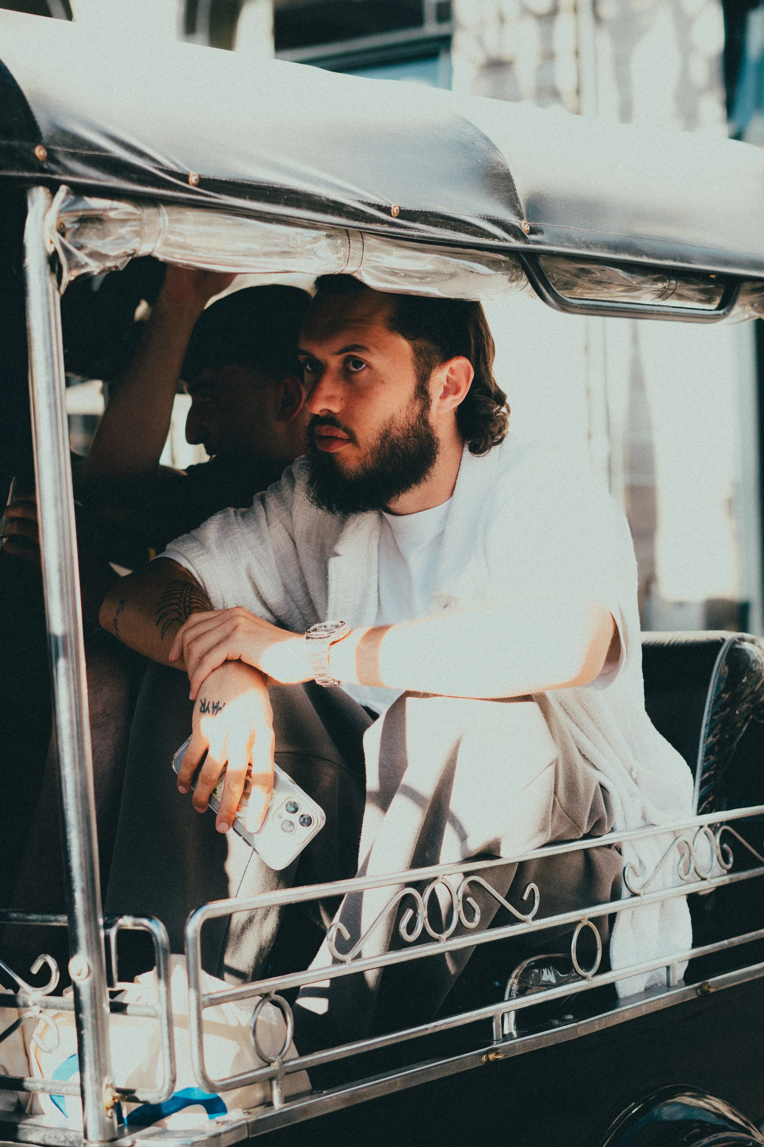 Un homme avec une barbe, portant un bracelet en métal, assis dans une charrette électrique, tenant un téléphone portable blanc, regardant pensivement vers l'avant.