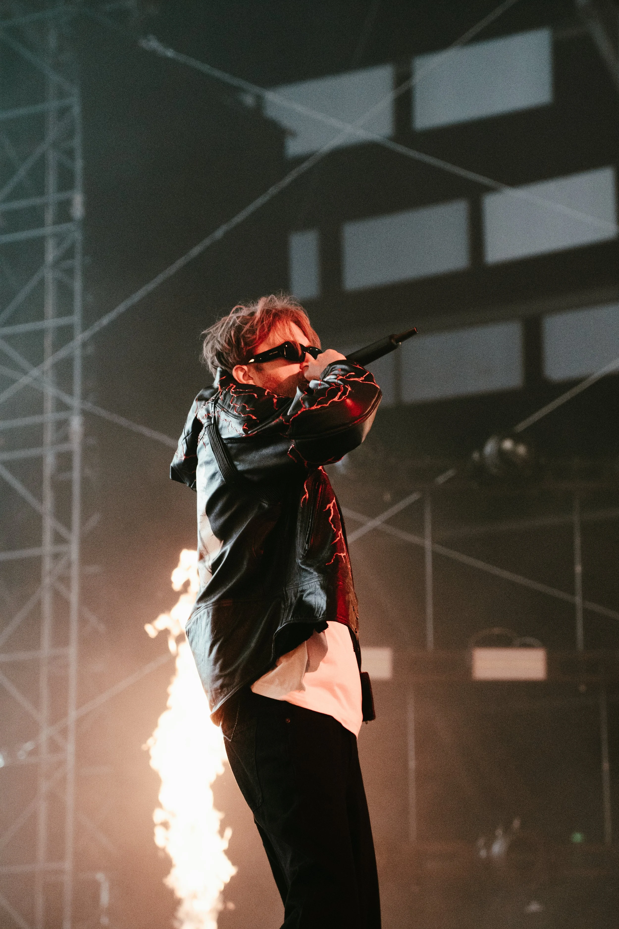 Un artiste en concert, portant des lunettes de soleil noires, un manteau en cuir noir avec des lignes rouges, chantant dans un microphone, avec des flammes en arrière-plan et une scène bardée de structures métalliques.