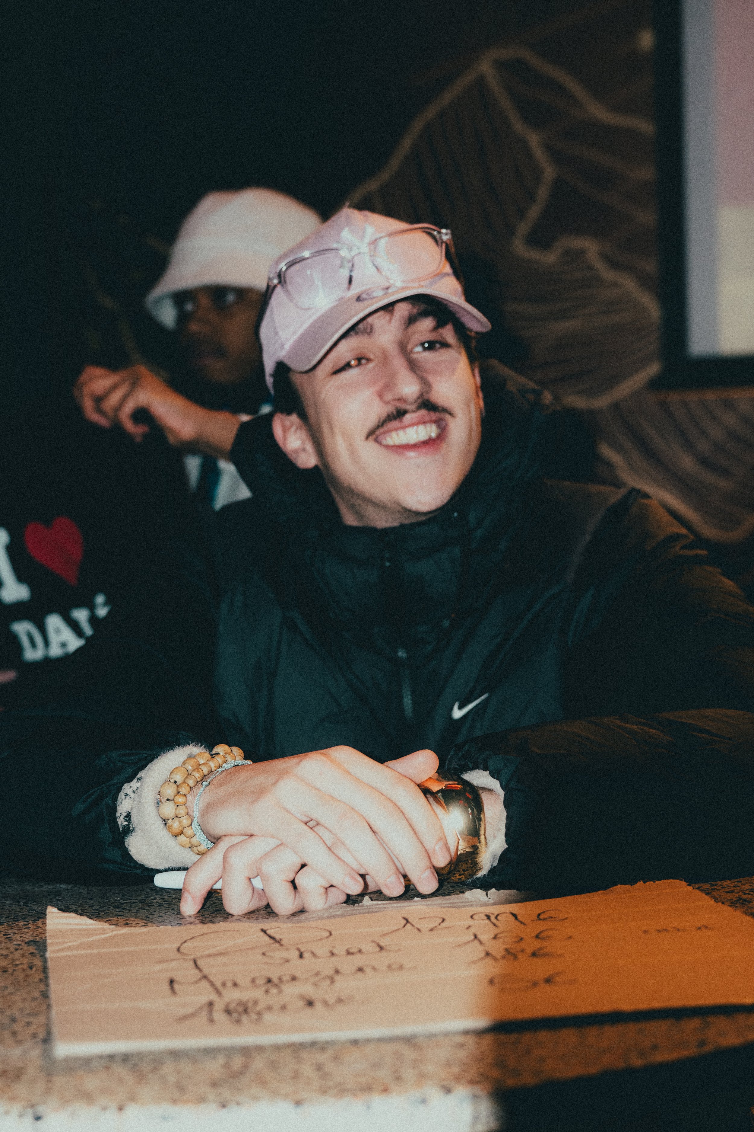 Une jeune homme souriant, portant une casquette rose et des lunettes, assis à une table avec un panneau en papier