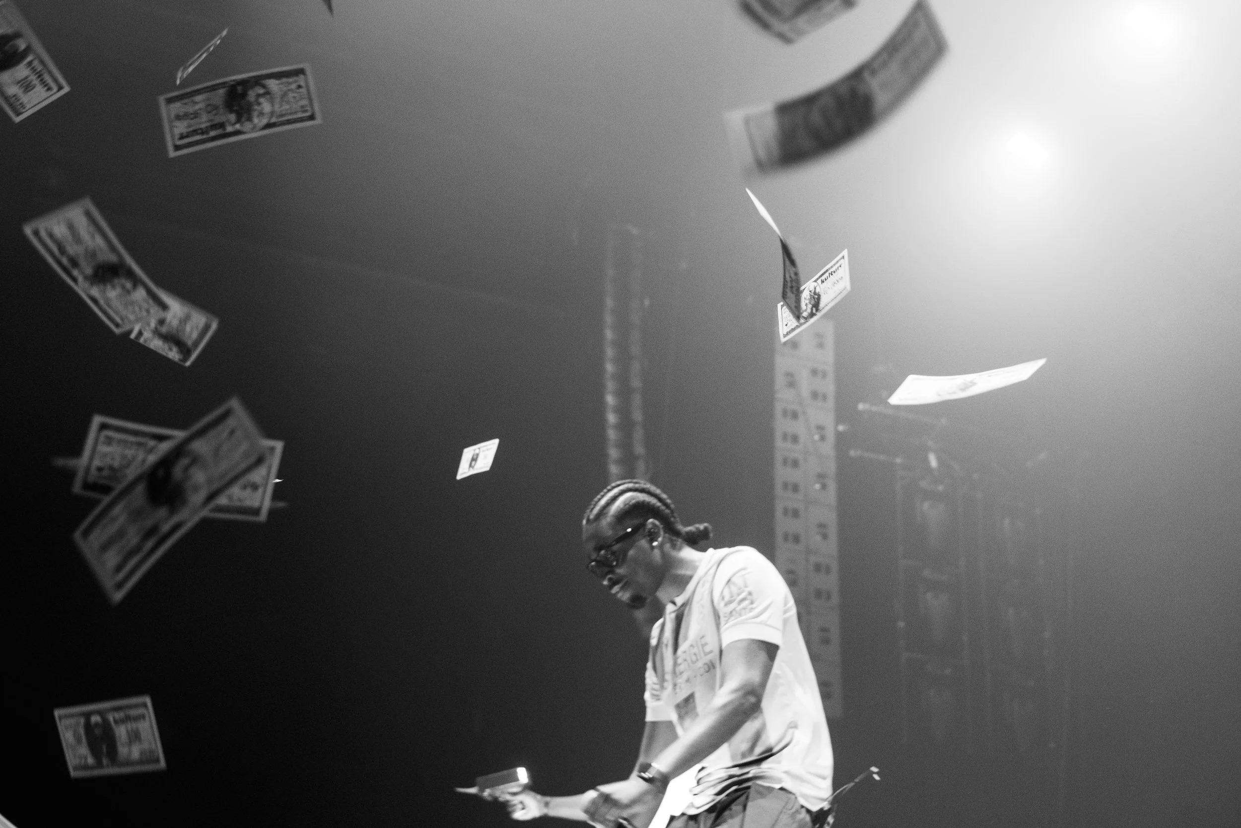 Une personne cheveux tressés portant des lunettes et une chemise blanche, debout sous une pluie de billets de banque, dans un environnement sombre et éclairé, semblant être sur scène ou dans un lieu de spectacle.