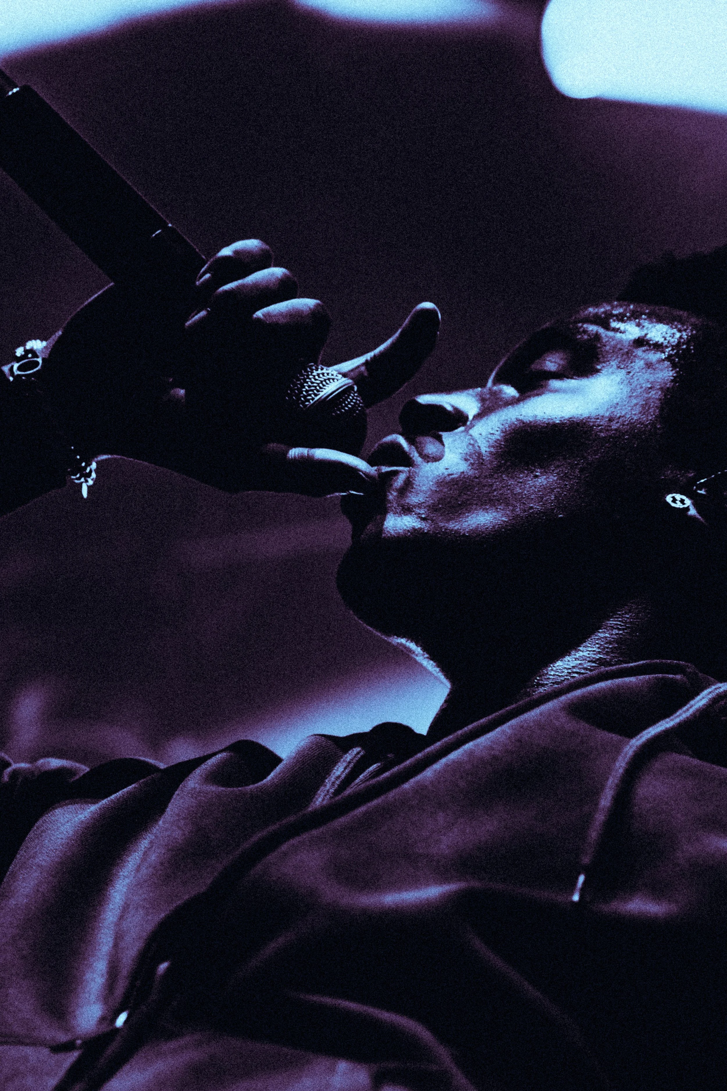 Un jeune homme avec des boucles d'oreilles porte un hoodie en cuir et tient un microphone, faisant un geste artistique avec sa main dans une ambiance sombre avec un éclairage violet.