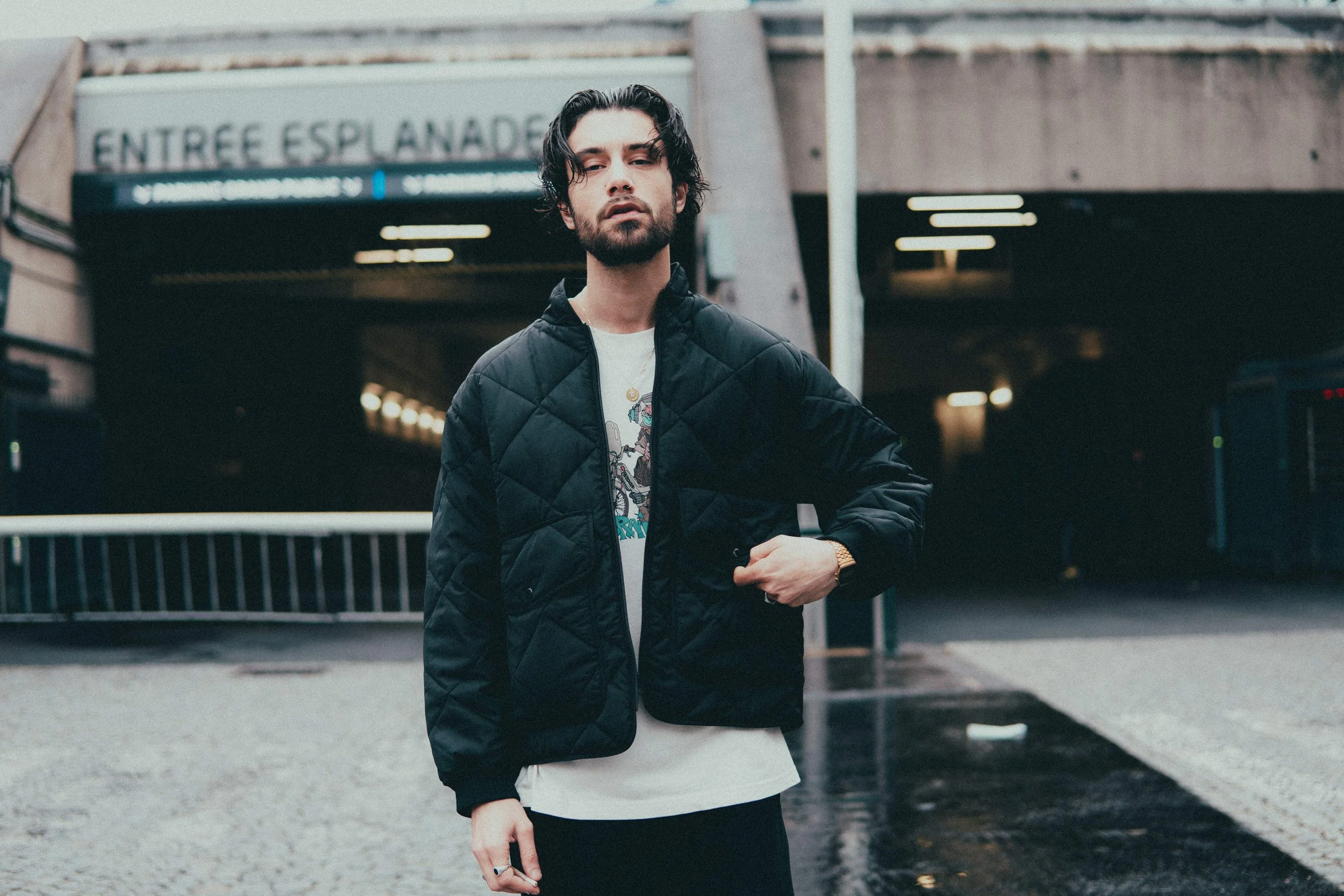 Un homme avec des cheveux longs et une barbe, porte une veste noire en denim et un t-shirt blanc, se tient devant une station de métro.
