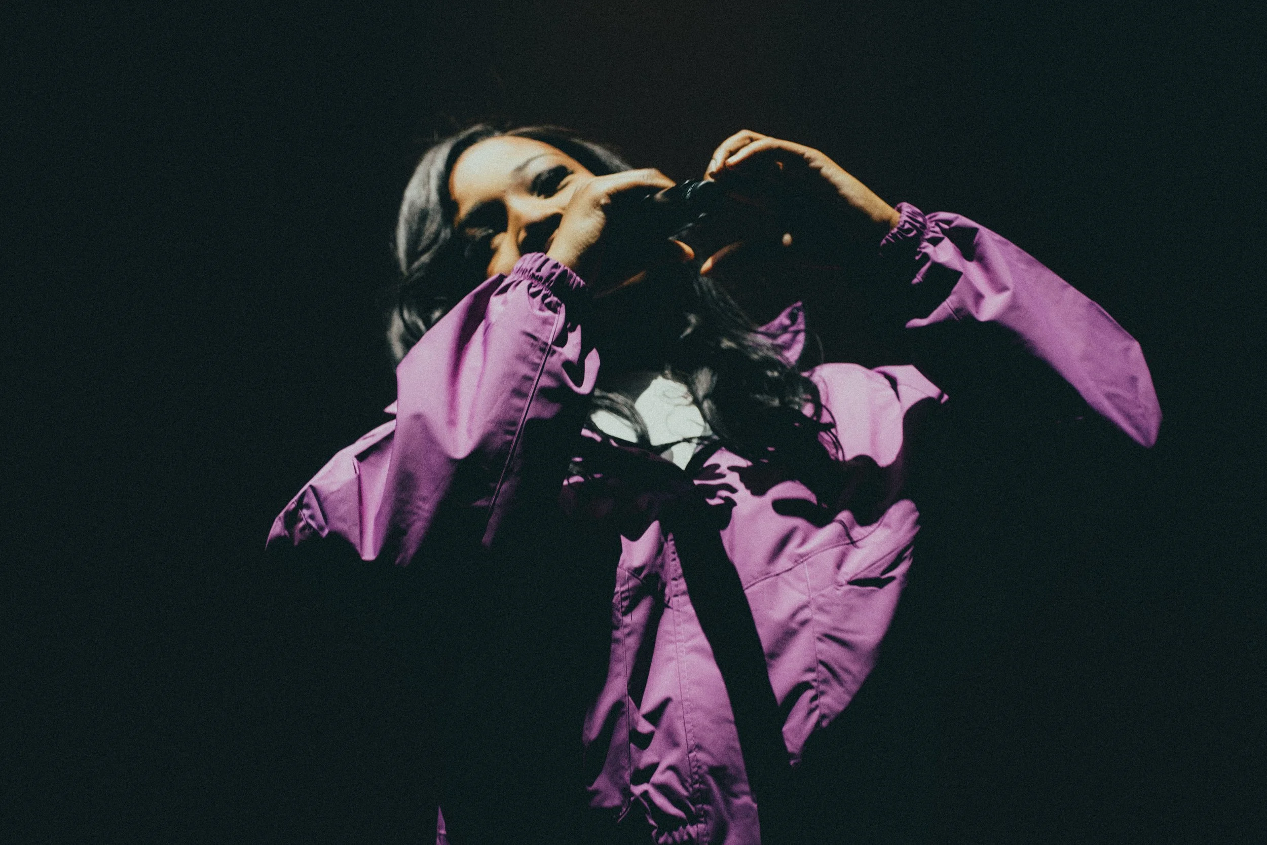 Une femme avec des cheveux longs foncés portant une veste violette, tenant un appareil photo, dans un environnement sombre.