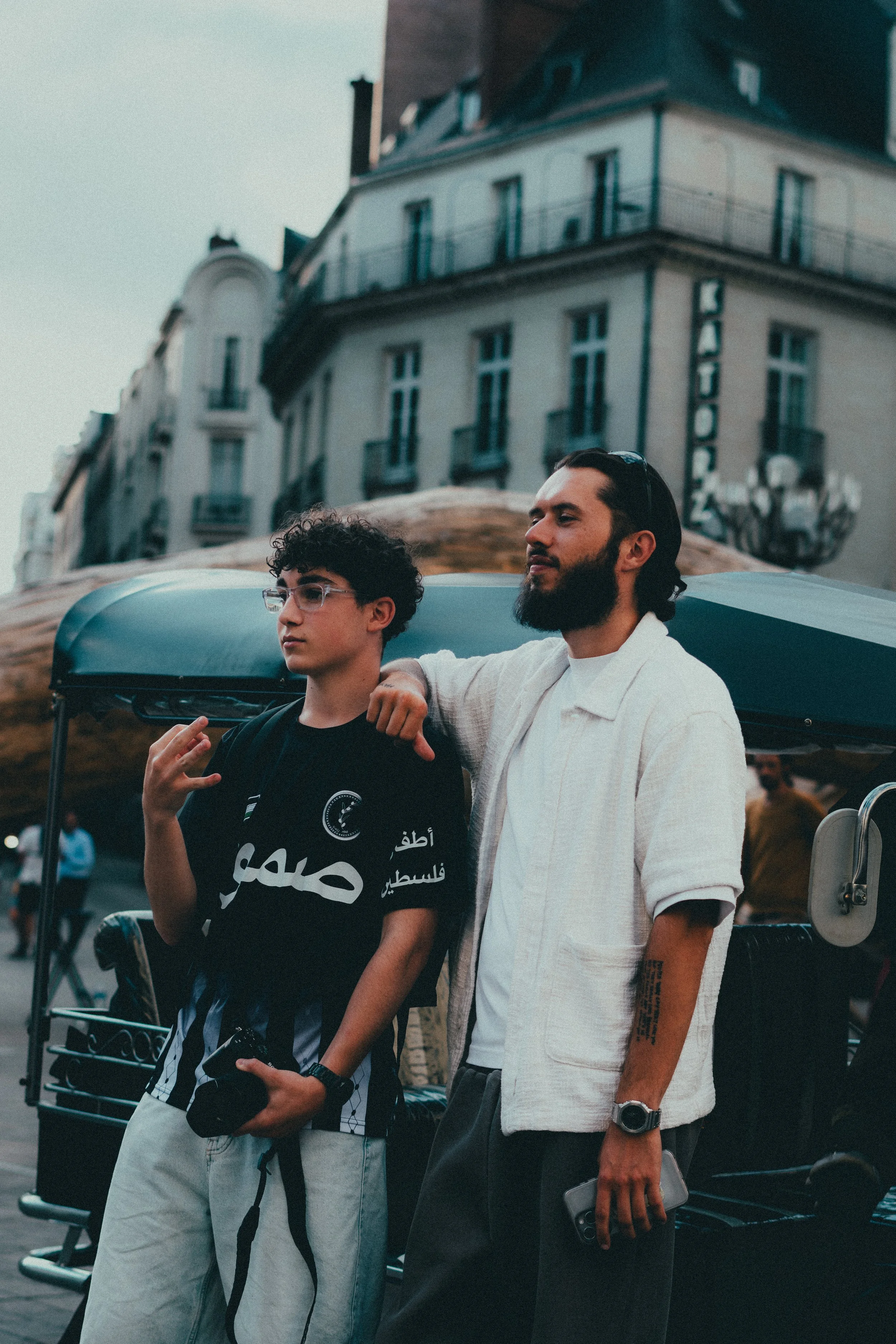 Deux jeunes hommes posent devant un tuk-tuk dans une rue urbaine. L'un porte un t-shirt noir avec des inscriptions en arabe, l'autre porte une chemise blanche ouverte.