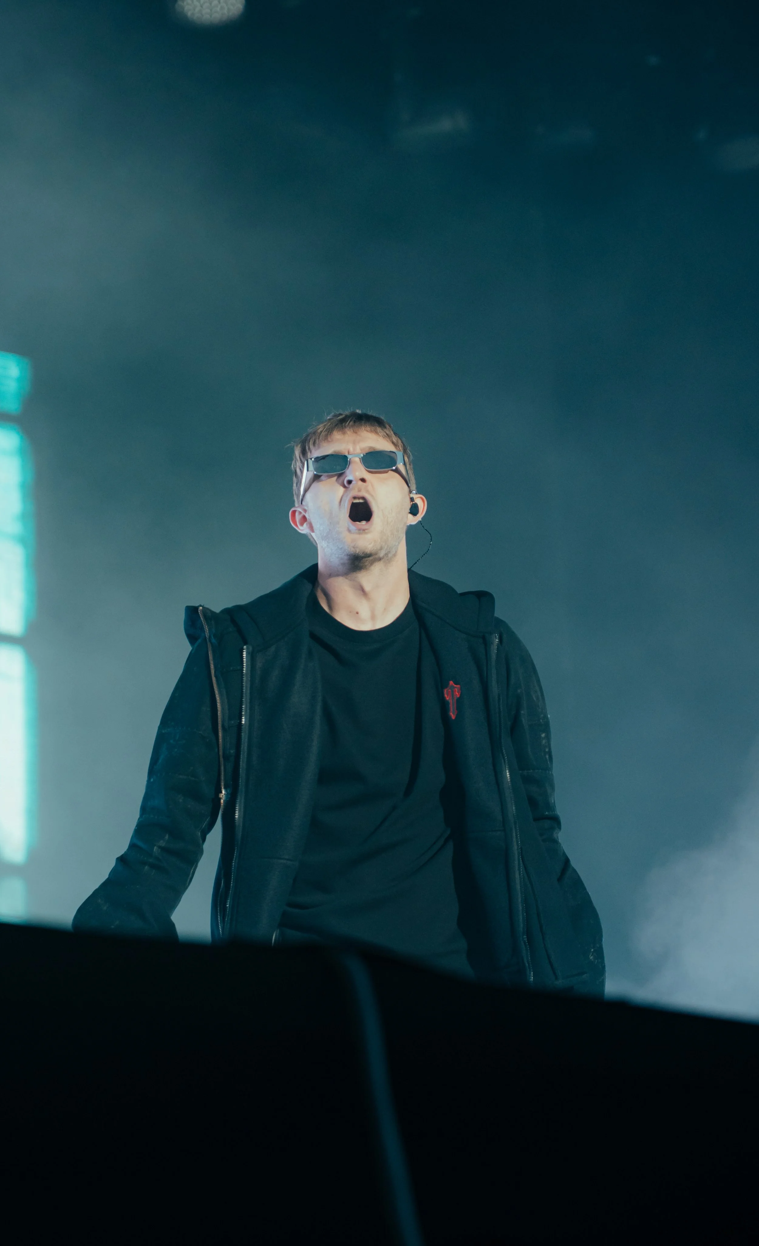 Un homme en veste noire et sunglasses chantant ou criant lors d'une performance sur scène.