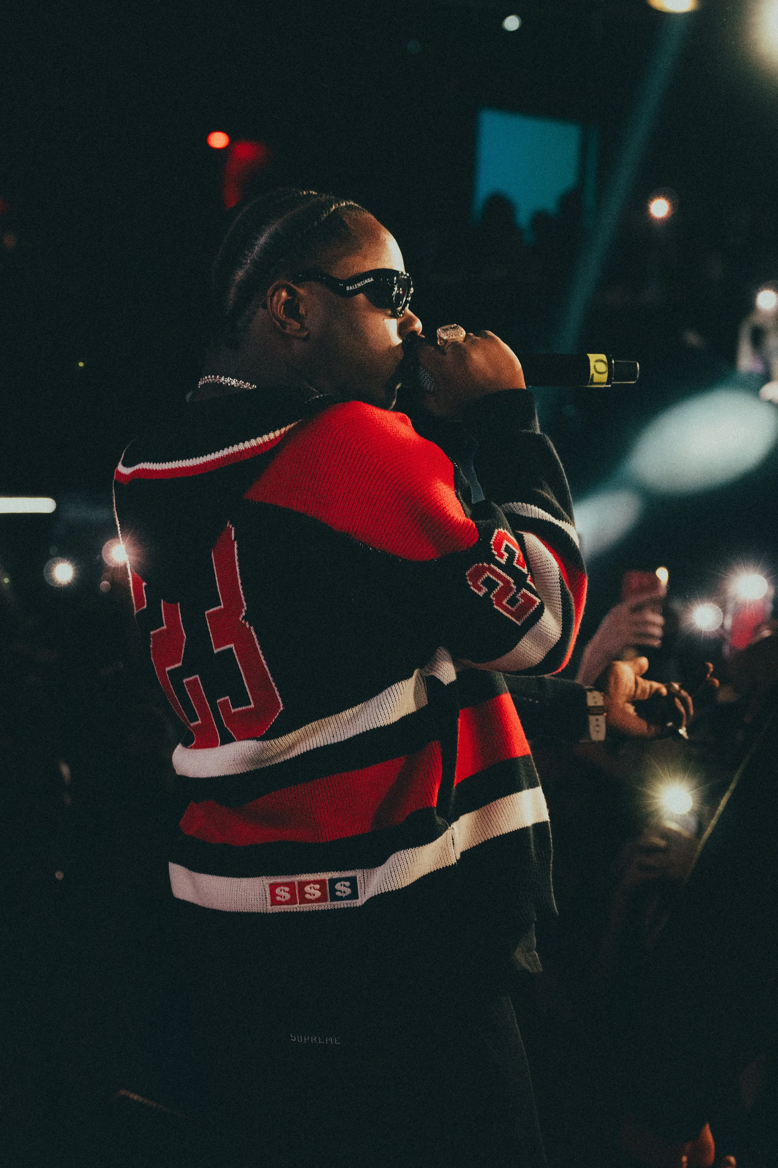 Un artiste portant une veste à rayures rouges, noires et blanches, et des lunettes de soleil, performe avec un micro dans une scène sombre avec des lumières et une foule en arrière-plan.