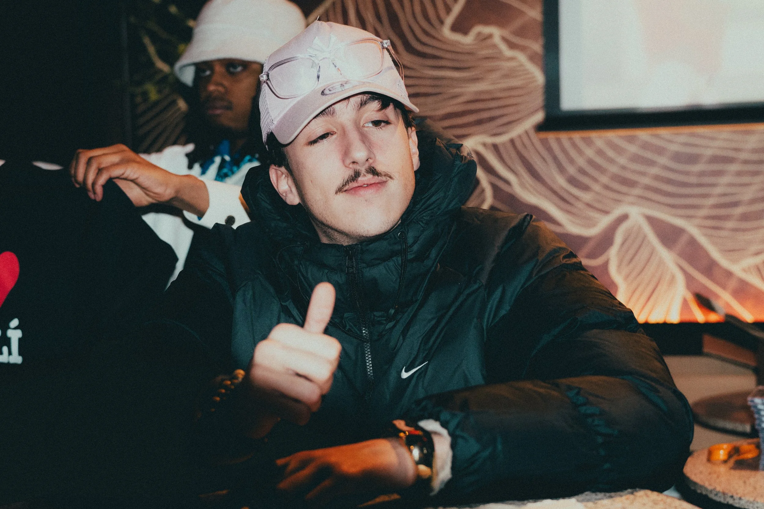 Un jeune homme portant une casquette blanc avec des lunettes sur le front, un manteau noir Nike, donne un pouce en l'air dans un intérieur avec un mur décoratif