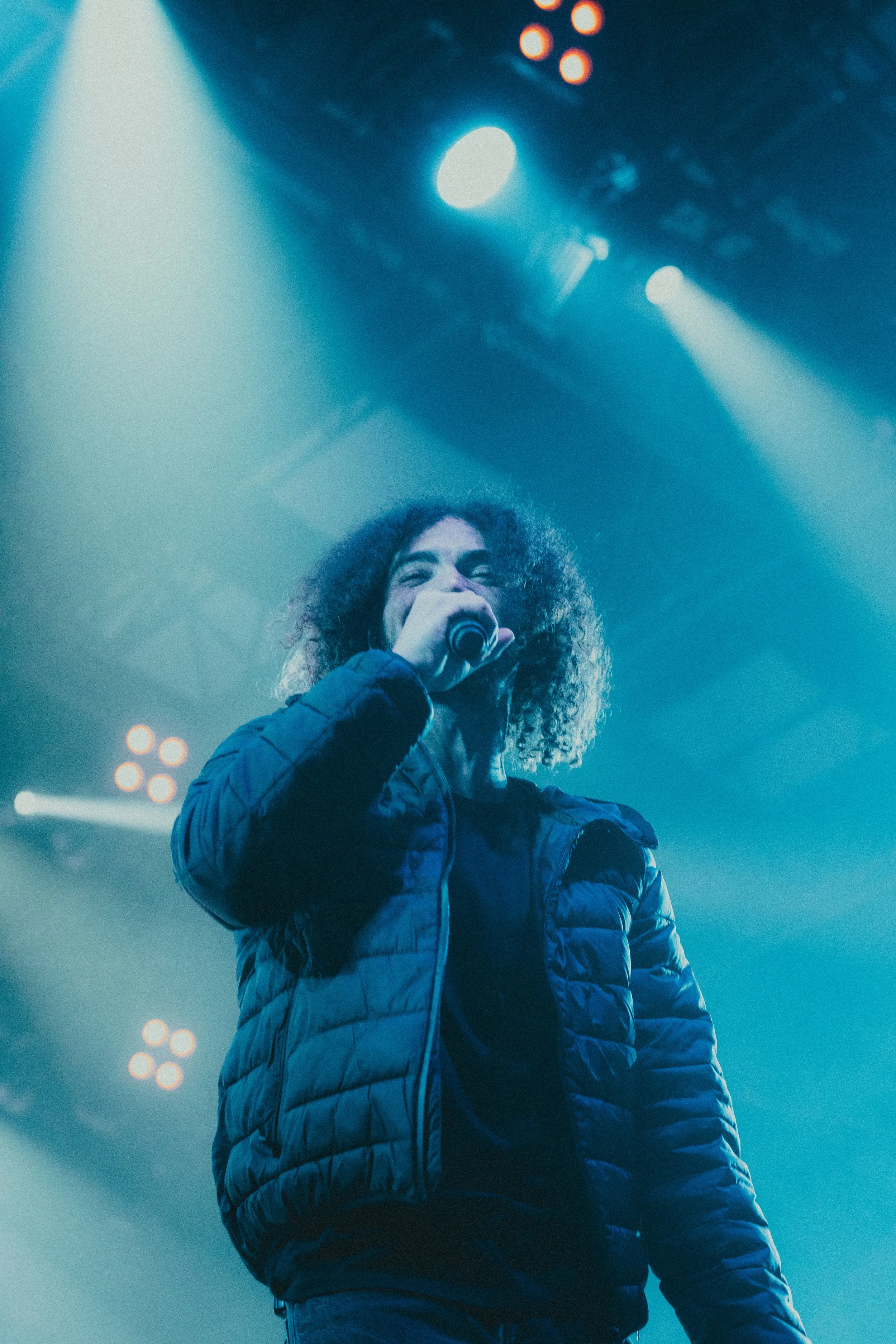 Une femme avec des cheveux bouclés tient un microphone sur scène, éclairée par des lumières de scène bleues et blanches.