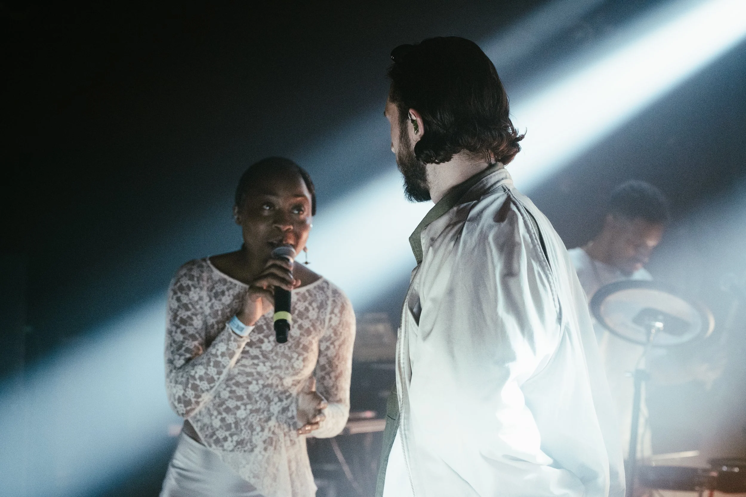 Un groupe de trois personnes en scène, avec une femme qui chante, un homme en face d'elle, et un musicien en arrière-plan.