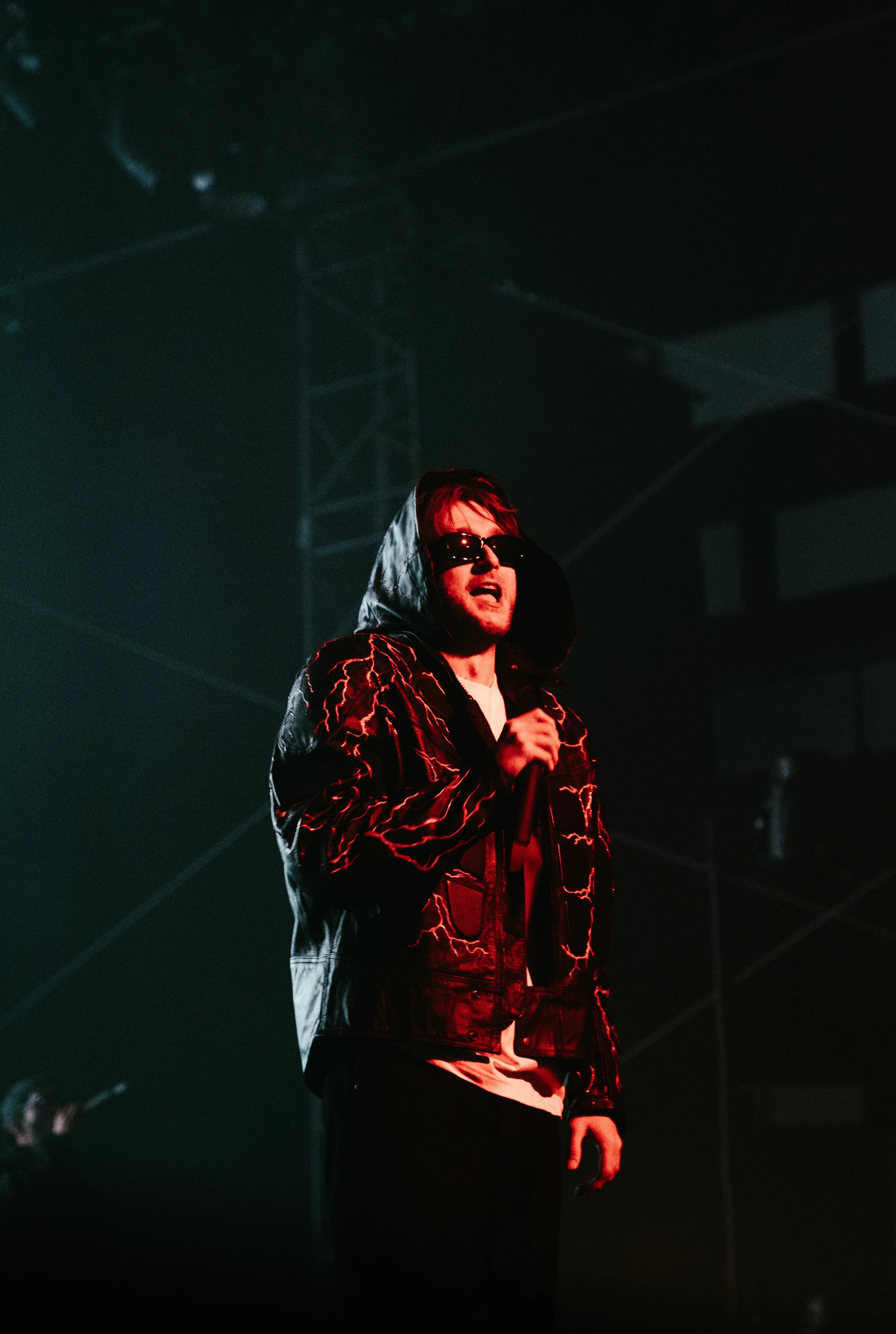 Un homme portant un sweat à capuche à capuche, des lunettes de soleil et tenant un microphone lors d'une performance sur scène, sous un éclairage tamisé.