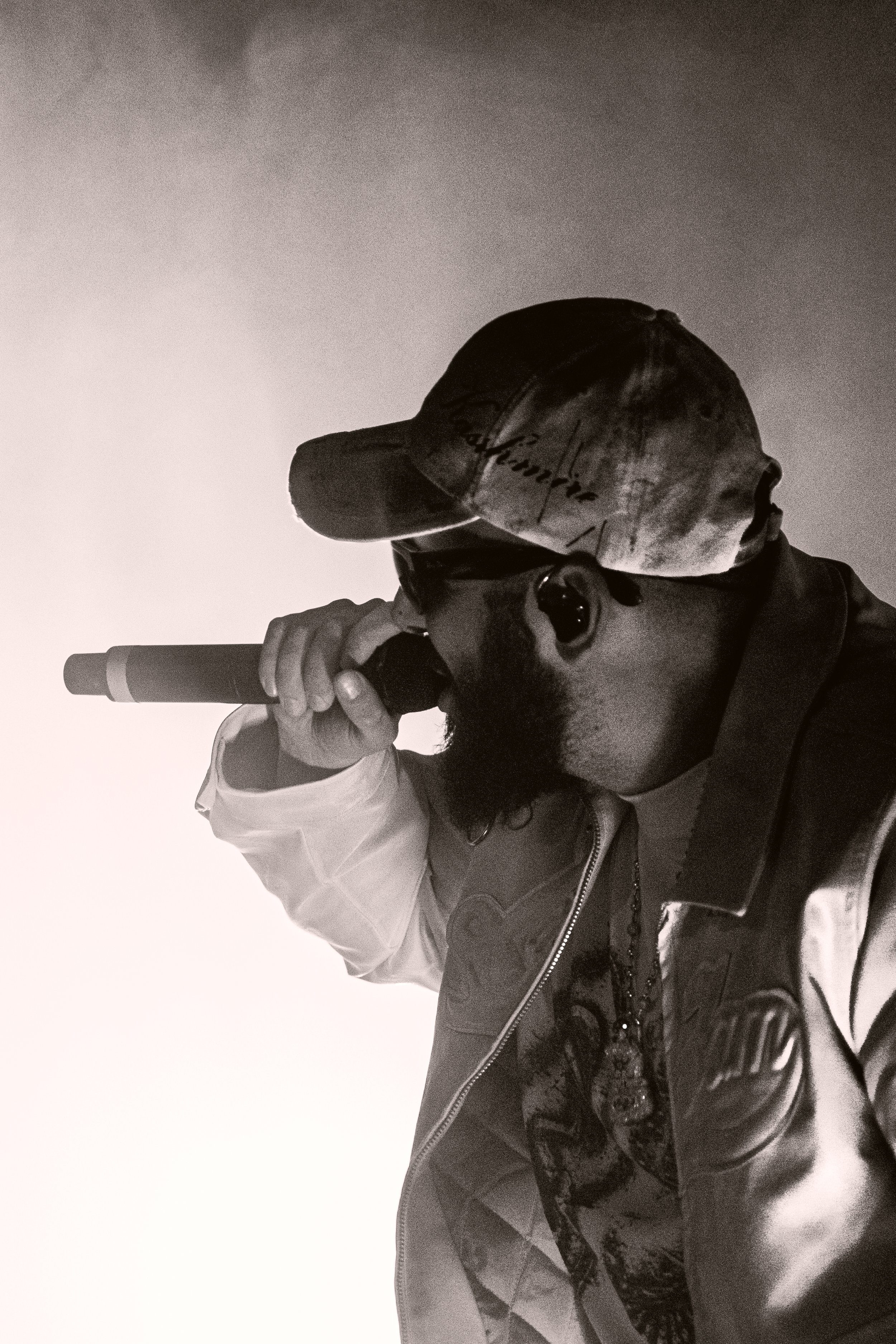 Un homme portant une casquette et une veste, tenant un microphone près de la bouche, en noir et blanc.