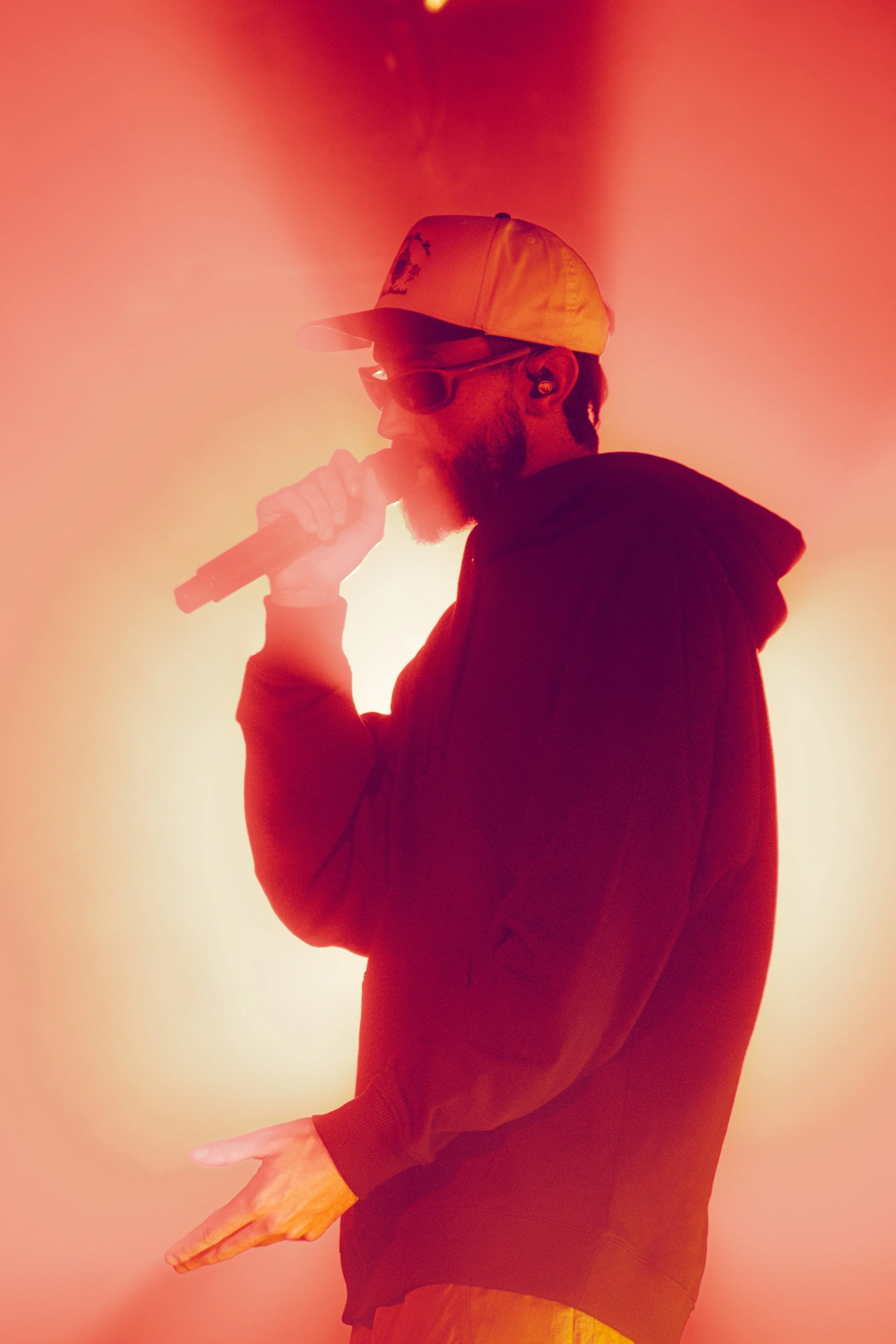 Un homme portant une casquette blanche, lunettes de soleil, tenant un microphone et chantant sur scène, avec un arrière-plan lumineux rouge/orangé.