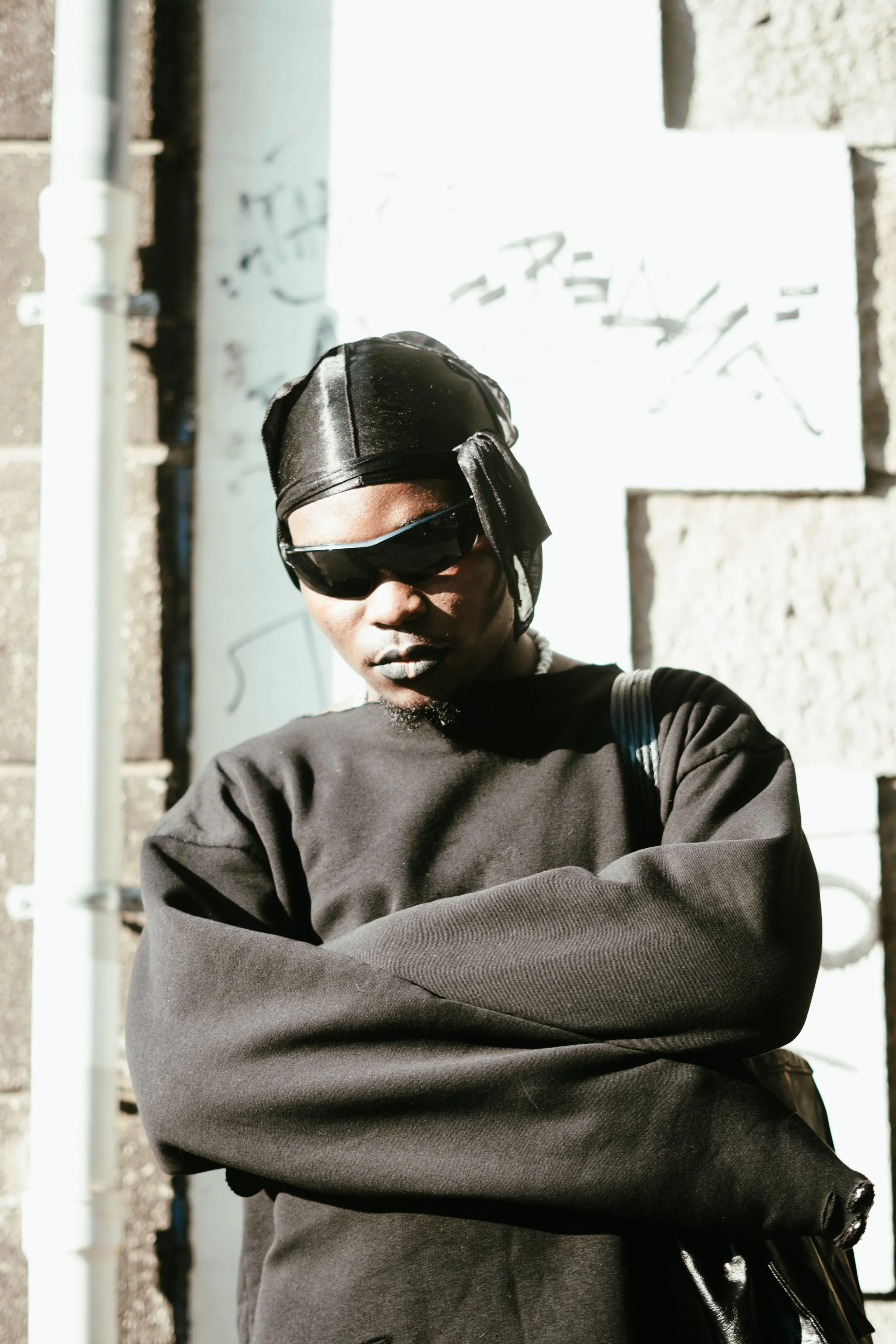 Un homme portant des lunettes de soleil sombres, un bandeau en satin noir, un sweat noir et un sac à dos, se tient contre un mur en pierre sous la lumière du soleil.