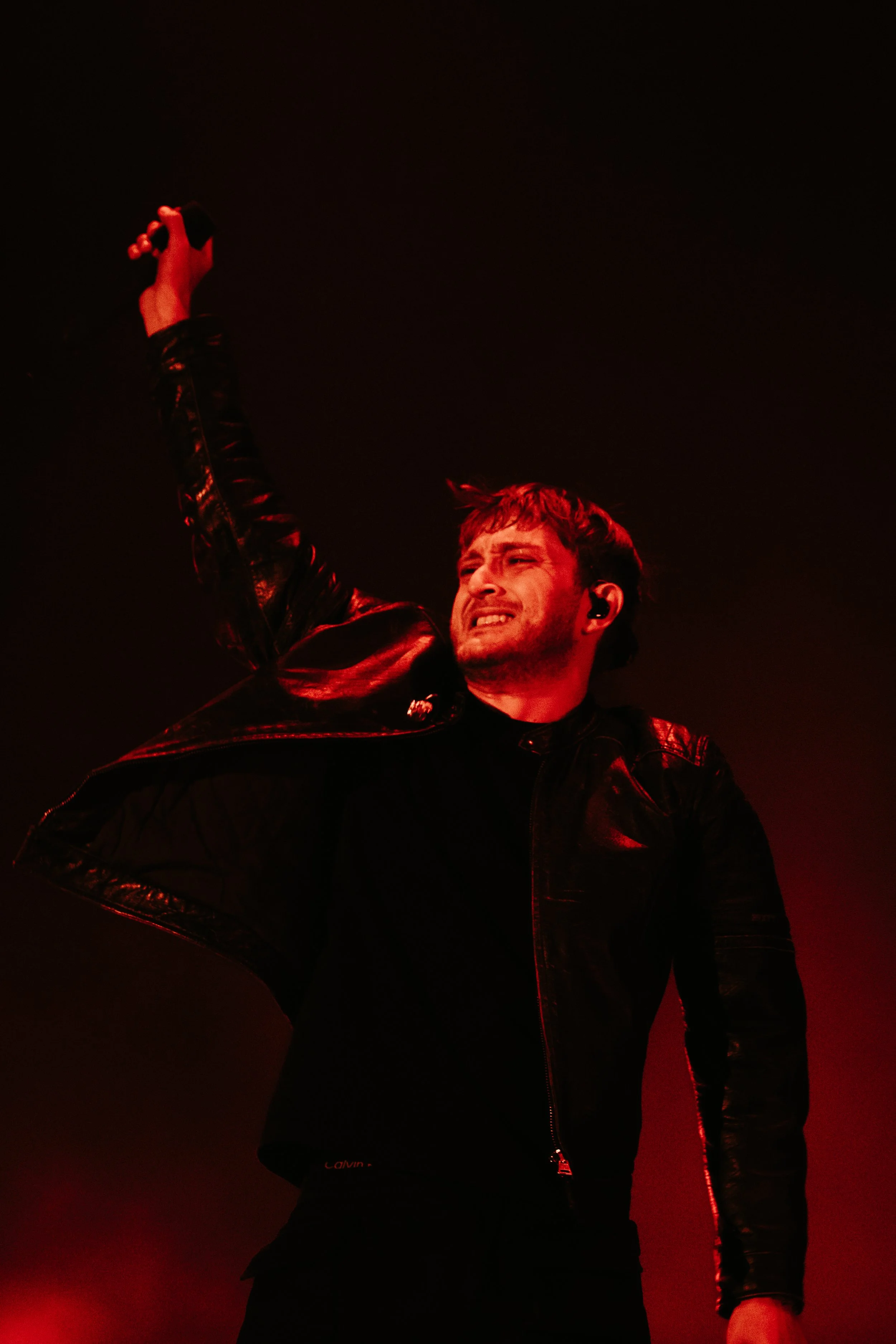 Un homme en pull noir et veste en cuire, tenant un microphone en l'air lors d'un concert, éclairé par une lumière rouge