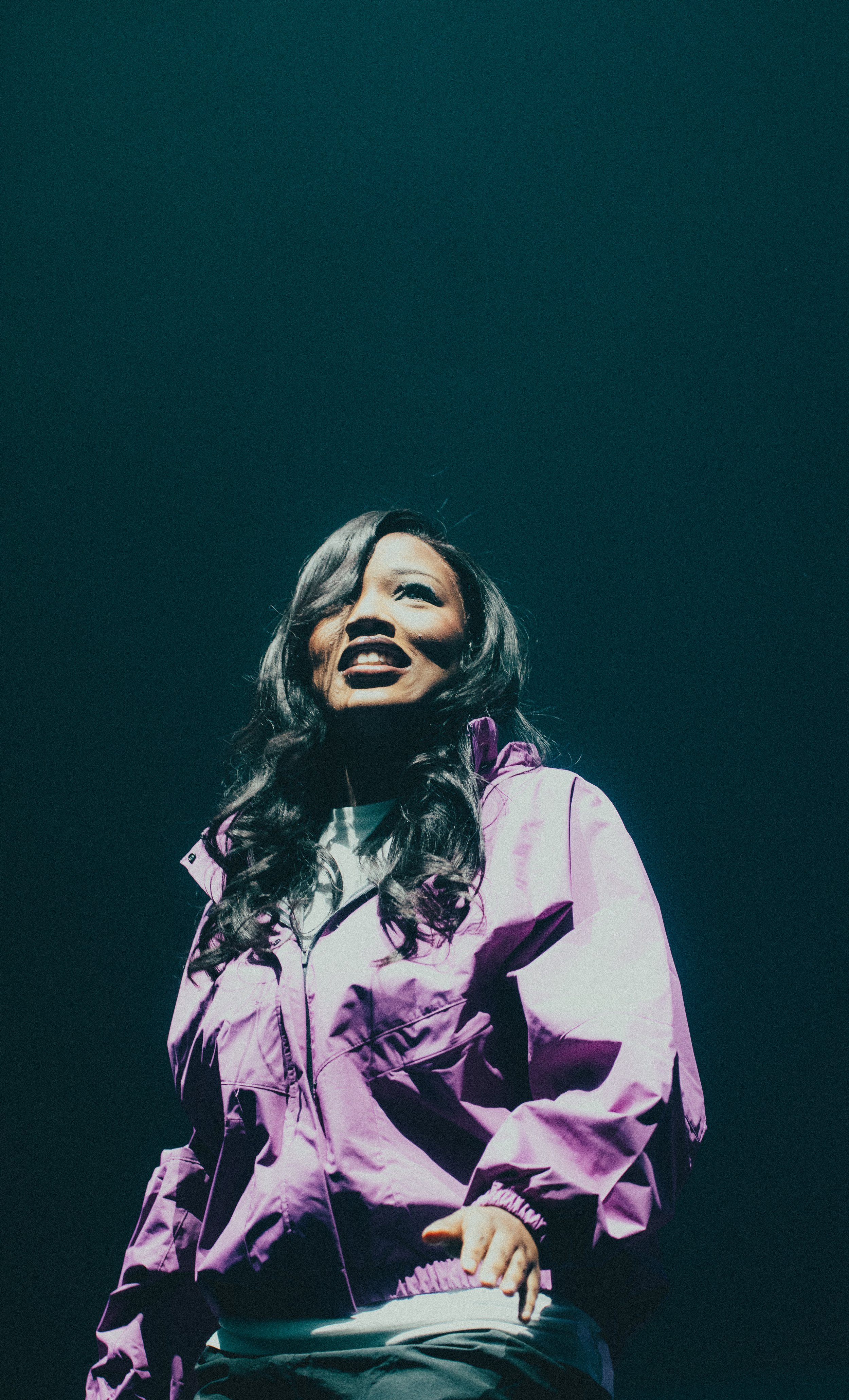 Une femme avec des cheveux longs et bouclés porte une veste violette, sourit et regarde vers le haut. Fond sombre.