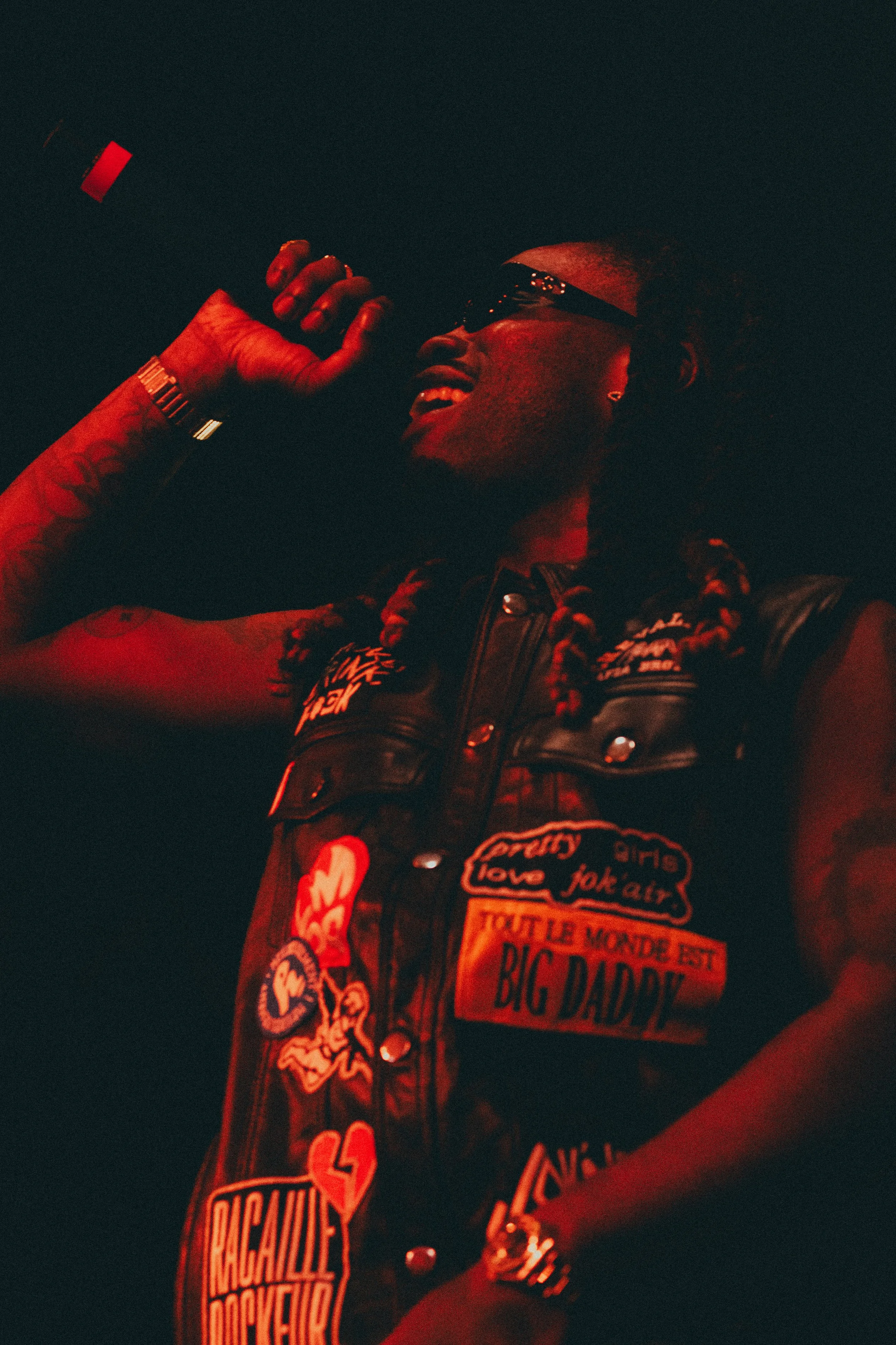 Une femme souriante porte des lunettes de soleil, une veste en cuir décorée de patchs colorés, et un bracelet en métal.