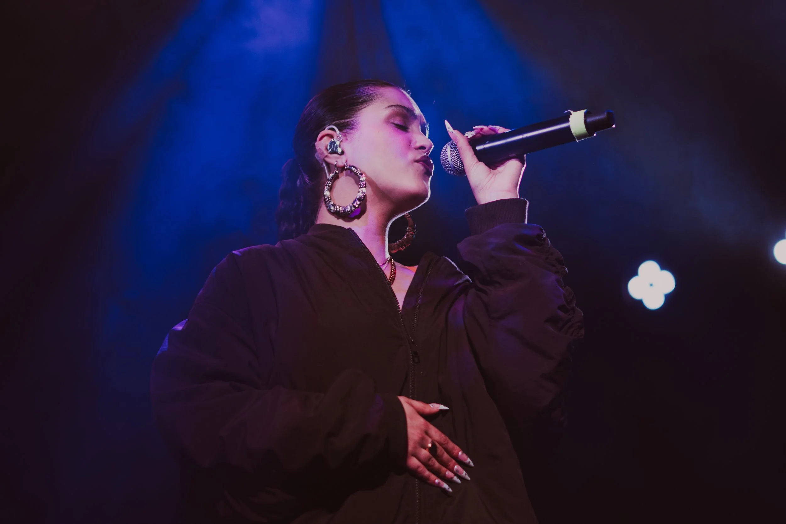 Une femme chante sur scène avec un micro, éclairée par des lumières violettes et bleues, portant de grosses boucles d'oreilles et une veste noire.