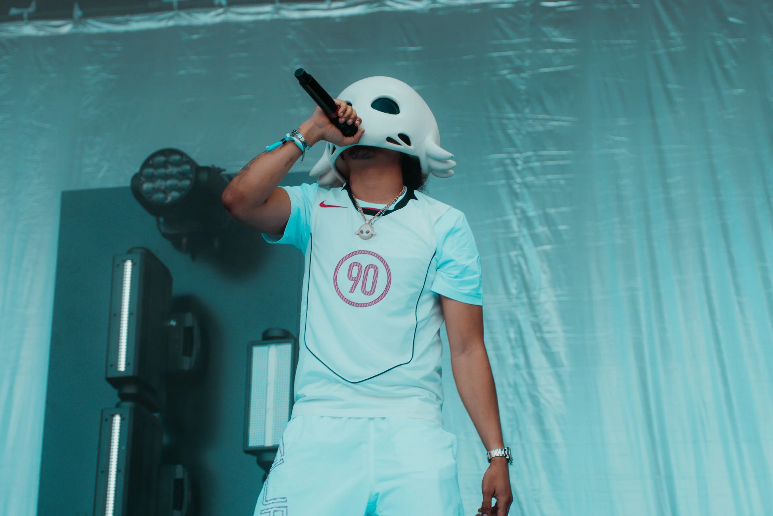 Personne portant un masque de tête d'animal, tenant un microphone lors d'une performance, vêtu d'un maillot blanc avec le numéro 90.