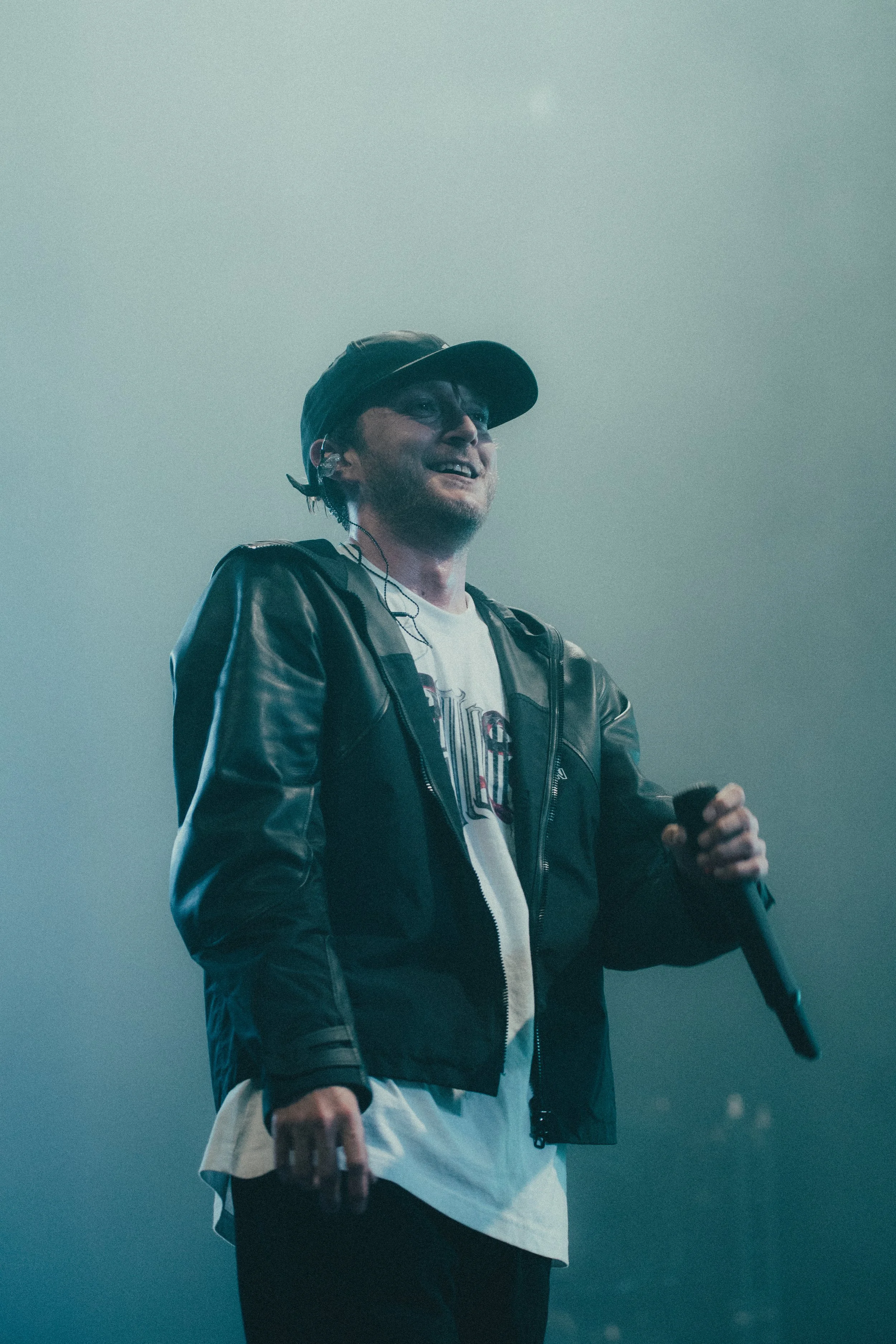 Un homme avec une casquette, portant une veste en cuir, tenant un microphone, en train de performer sur scène