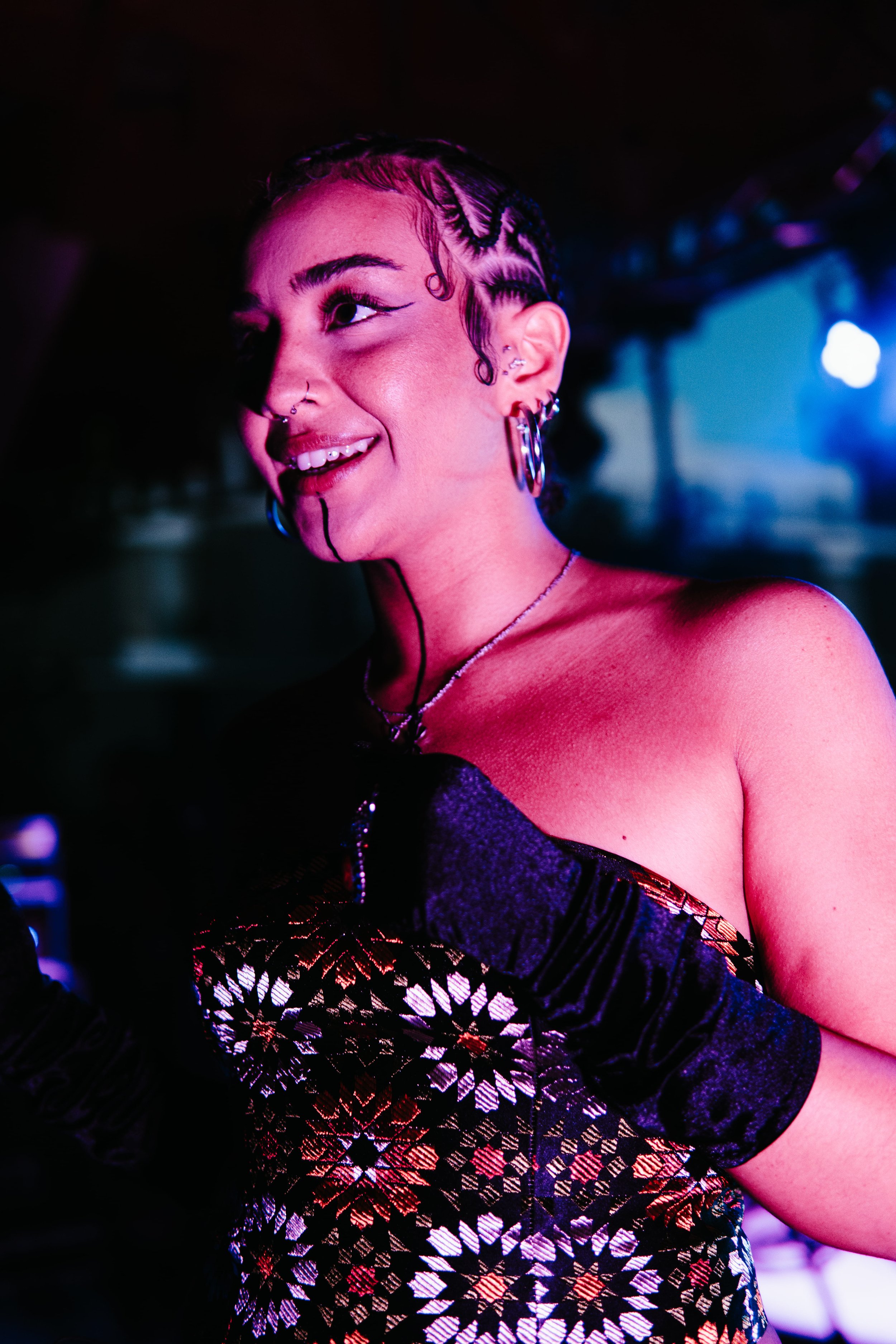 Jeune femme souriante avec des cheveux plats et des bouclettes, portant un vêtement à motifs colorés, portant des bijoux piercings et des maquillage artistique dans un environnement sombre avec des lumières vives.