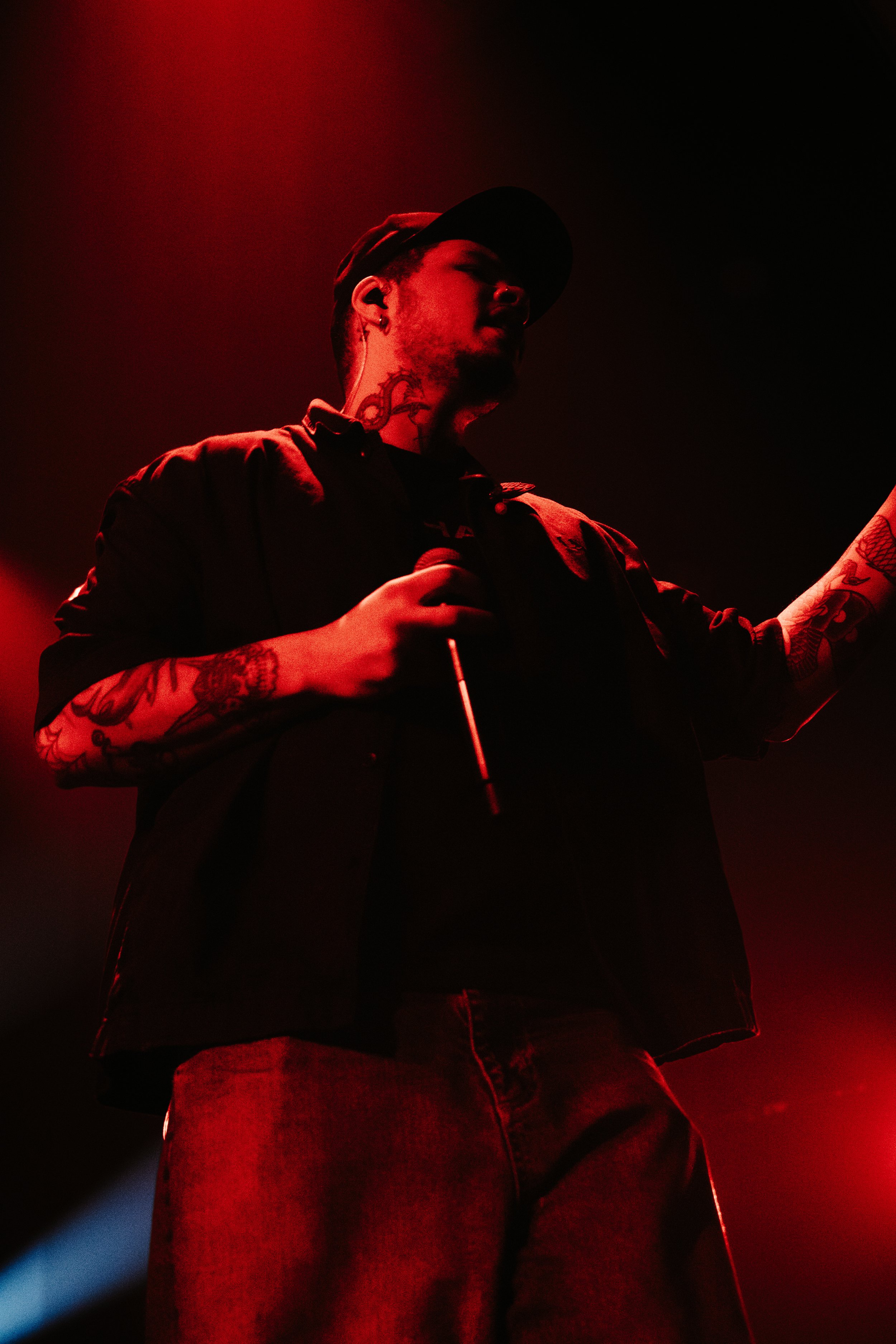 Un homme tatoué portant une casquette noire et une veste noire, tenant un microphone, sous un éclairage rouge.