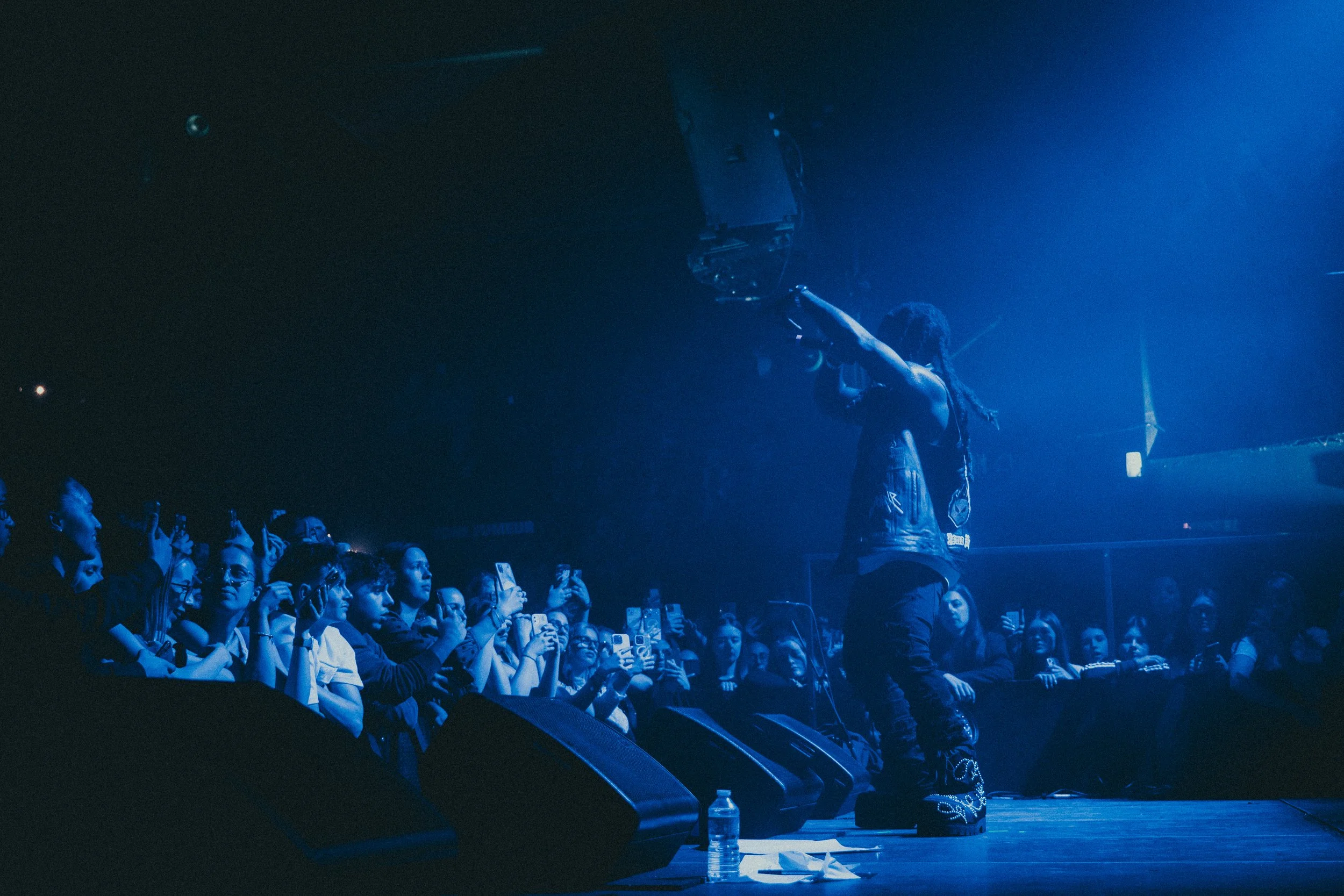 Un artiste avec des dreadlocks, portant des vêtements amples, performe sur scène devant un public nombreux qui prend des photos et filme avec leurs téléphones, dans des lumières bleues.