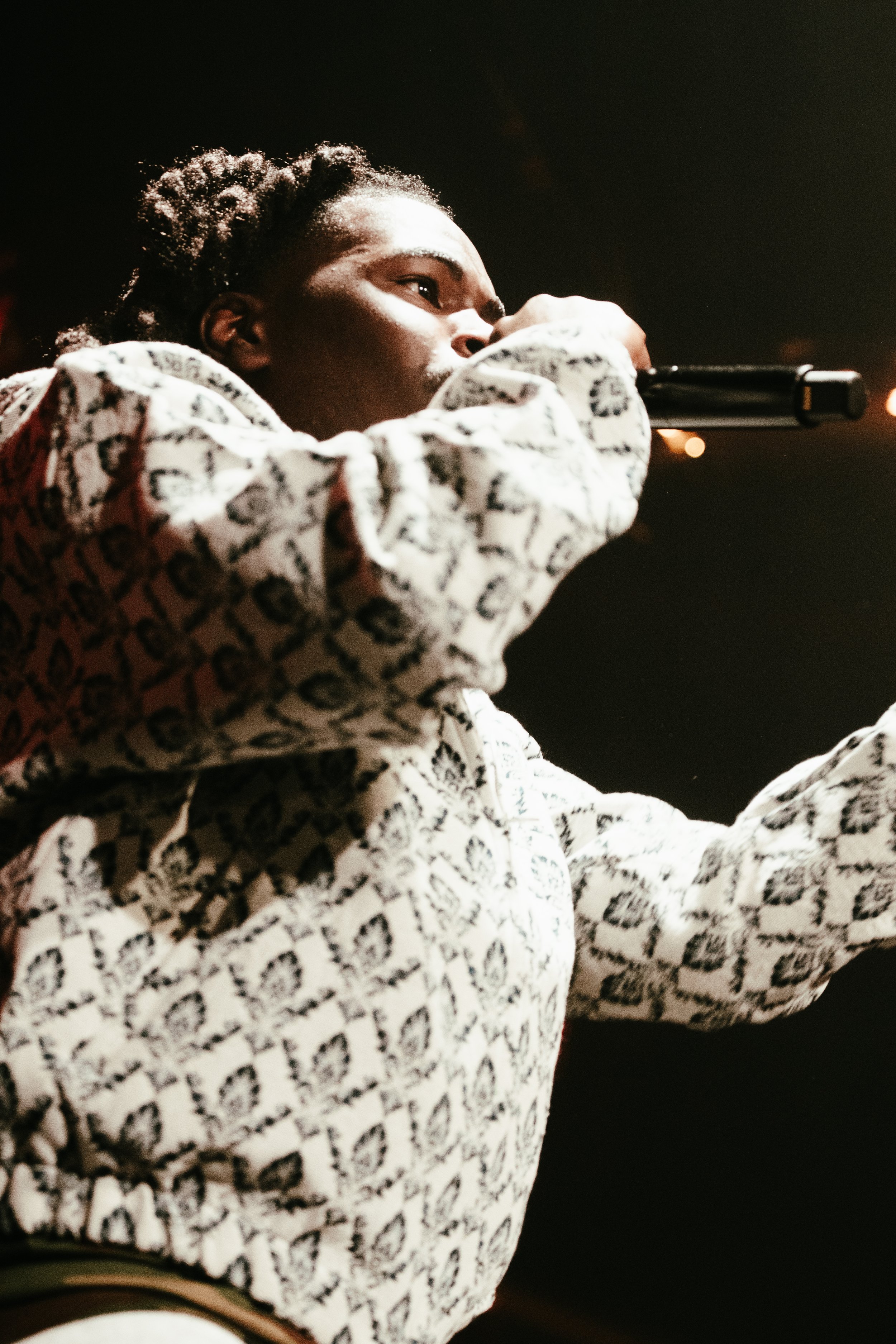 Un artiste ou chanteur noir portant un sweat à motif géométrique, tenant un micro et en train de performer sur scène.