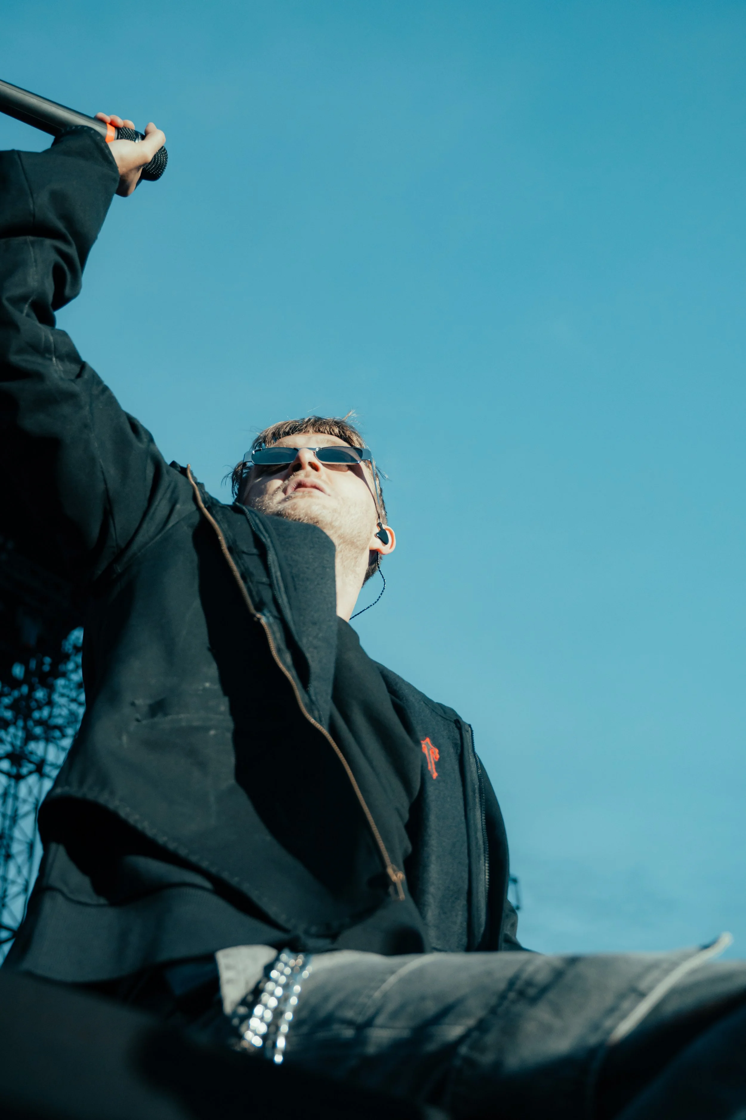 Un homme portant des lunettes de soleil, une veste noire et des écouteurs, tenant une canne ou un bâton, regardant vers le haut, avec un ciel bleu en arrière-plan.