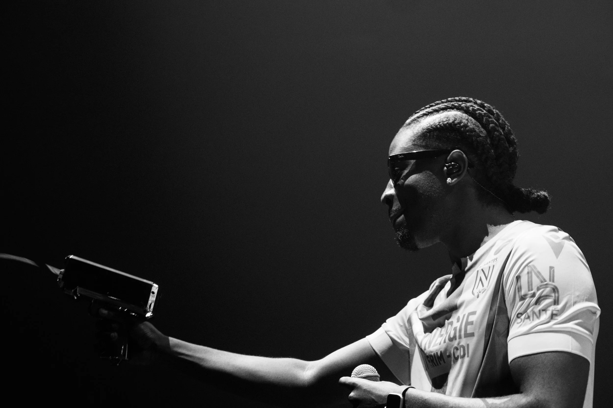 Un homme portant des lunettes de soleil tient un microphone dans une main et un objet dans l'autre, avec un fond sombre.