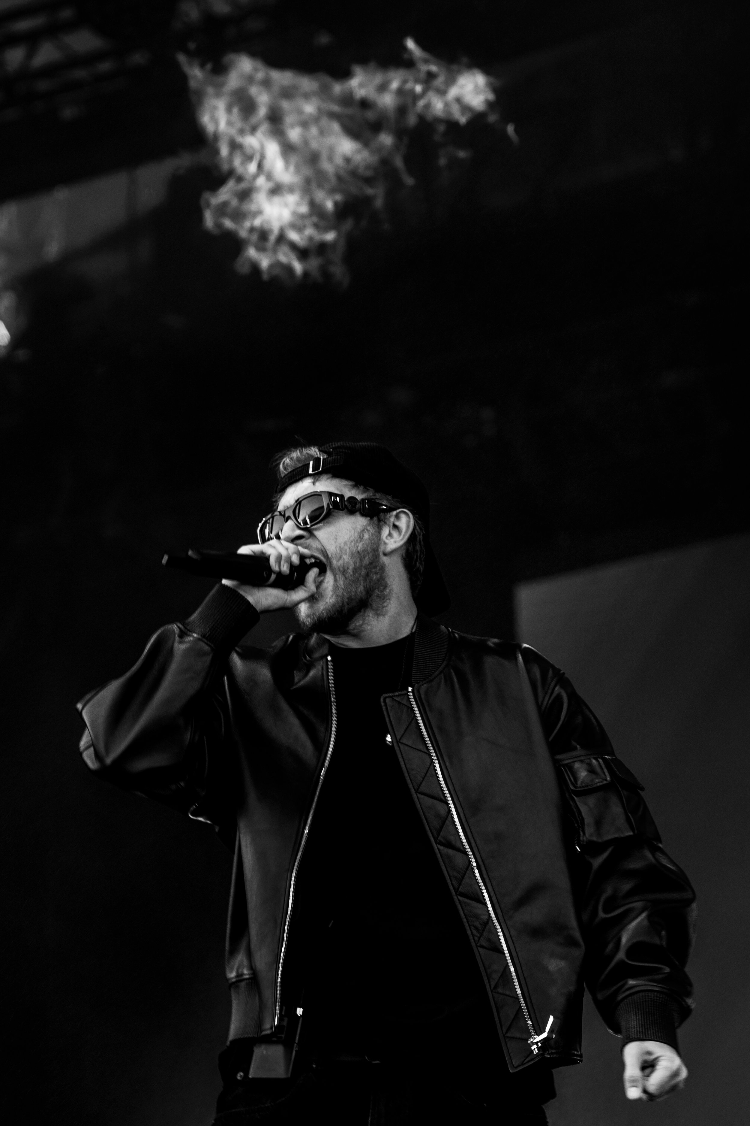 Un homme portant des lunettes de soleil, une casquette à l'envers, un blouson en cuir noir, tenant un micro et chantant sur scène.