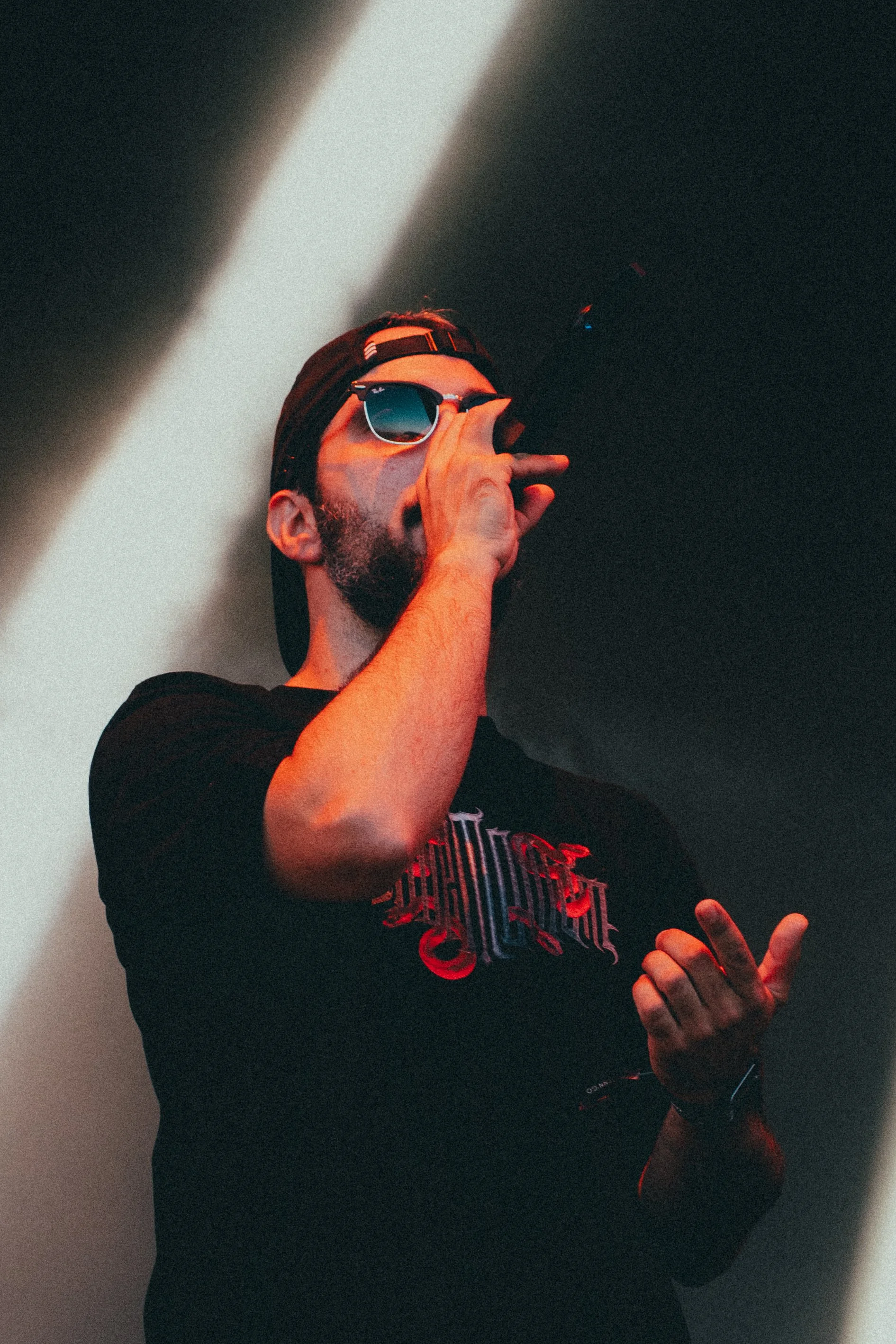 Un homme portant des lunettes de soleil, un t-shirt noir à motif rouge, une casquette à l'envers, tenant un micro, en train de chanter ou de parler, sur fond sombre avec un rayon lumineux.