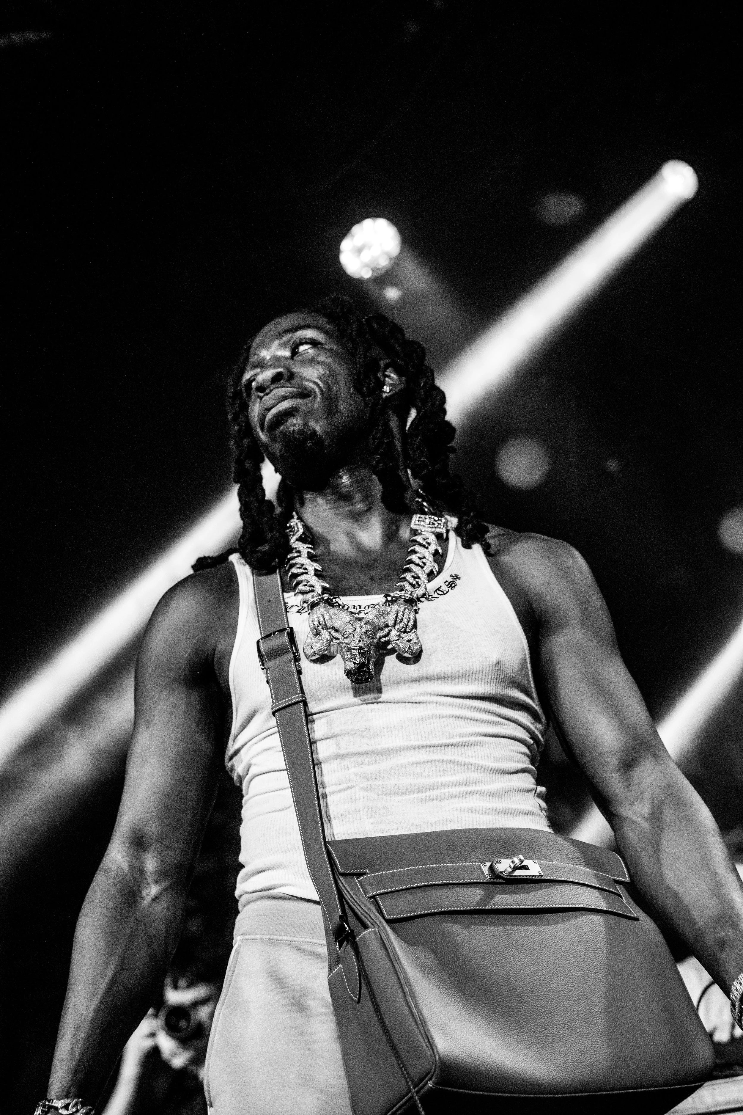 Un homme avec des dreadlocks, portant un t-shirt sans manches, un collier avec des pendentifs, et un sac à bandoulière, sur scène avec des éclairages.