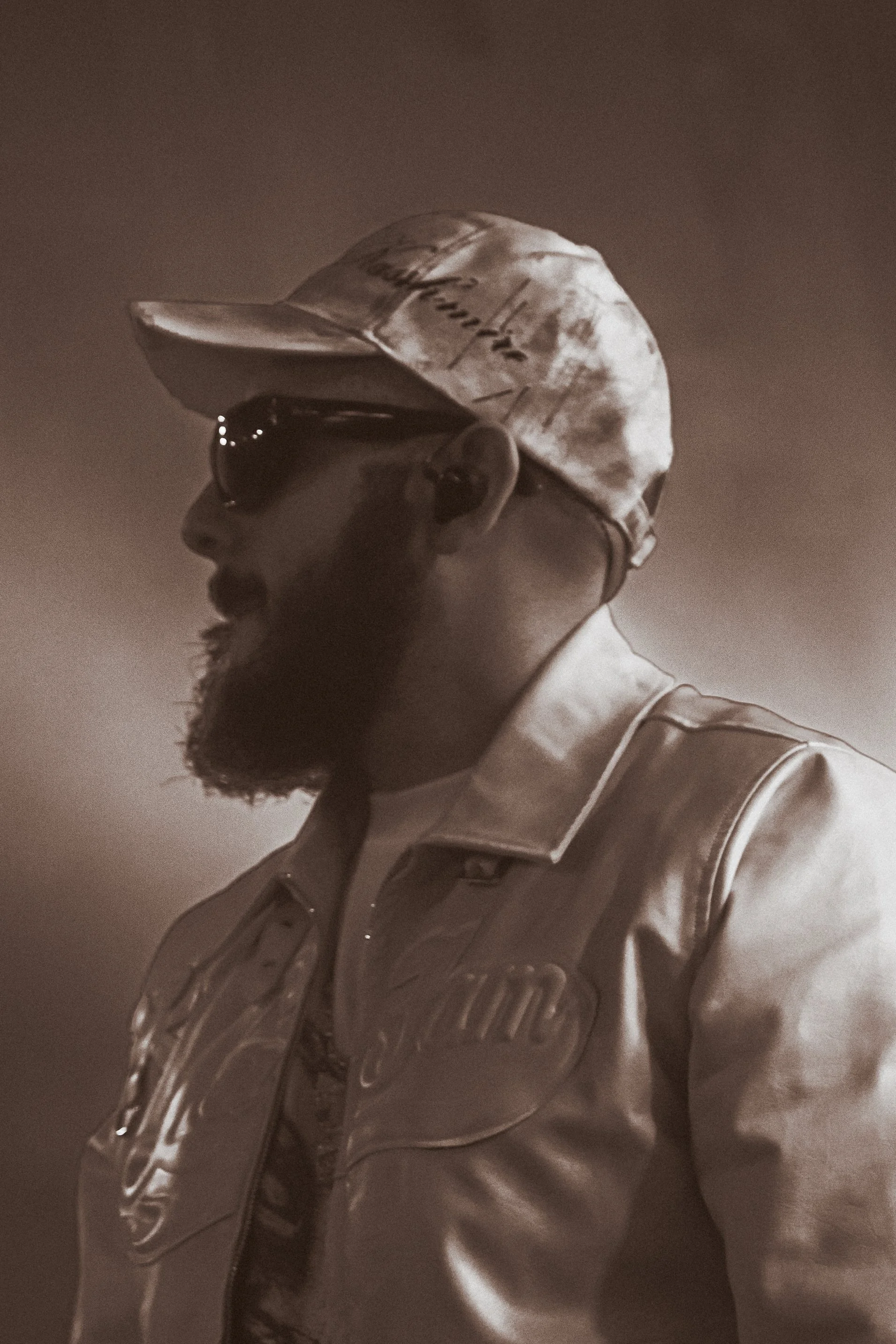 Portrait d'un homme portant une casquette, des lunettes de soleil et une veste en cuir, avec une barbe et un sérieux élégant.