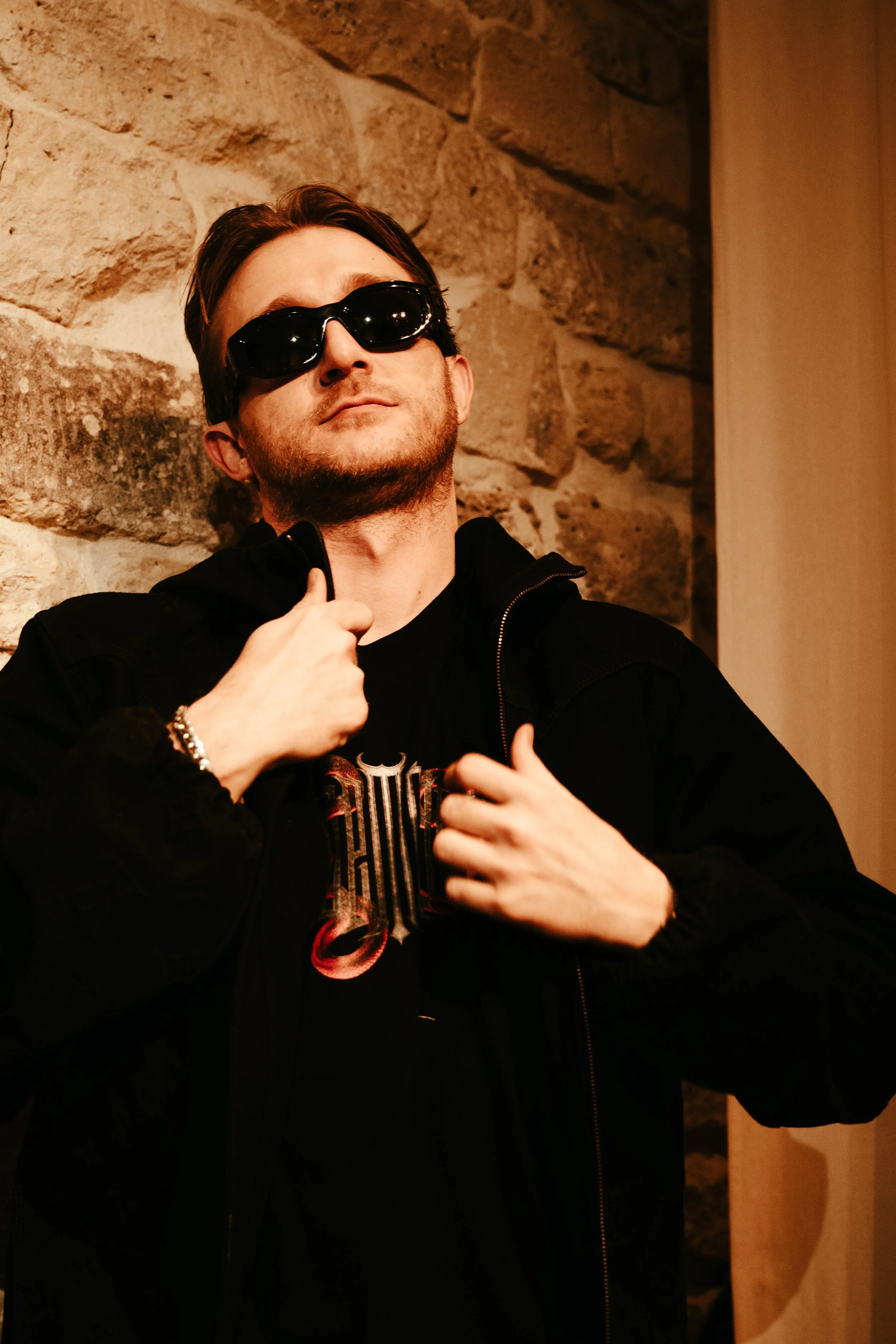 Un homme portant des lunettes de soleil noires, une veste noire, et un t-shirt noir avec un design métallisé, pose contre un mur en pierre, avec une attitude confiante.