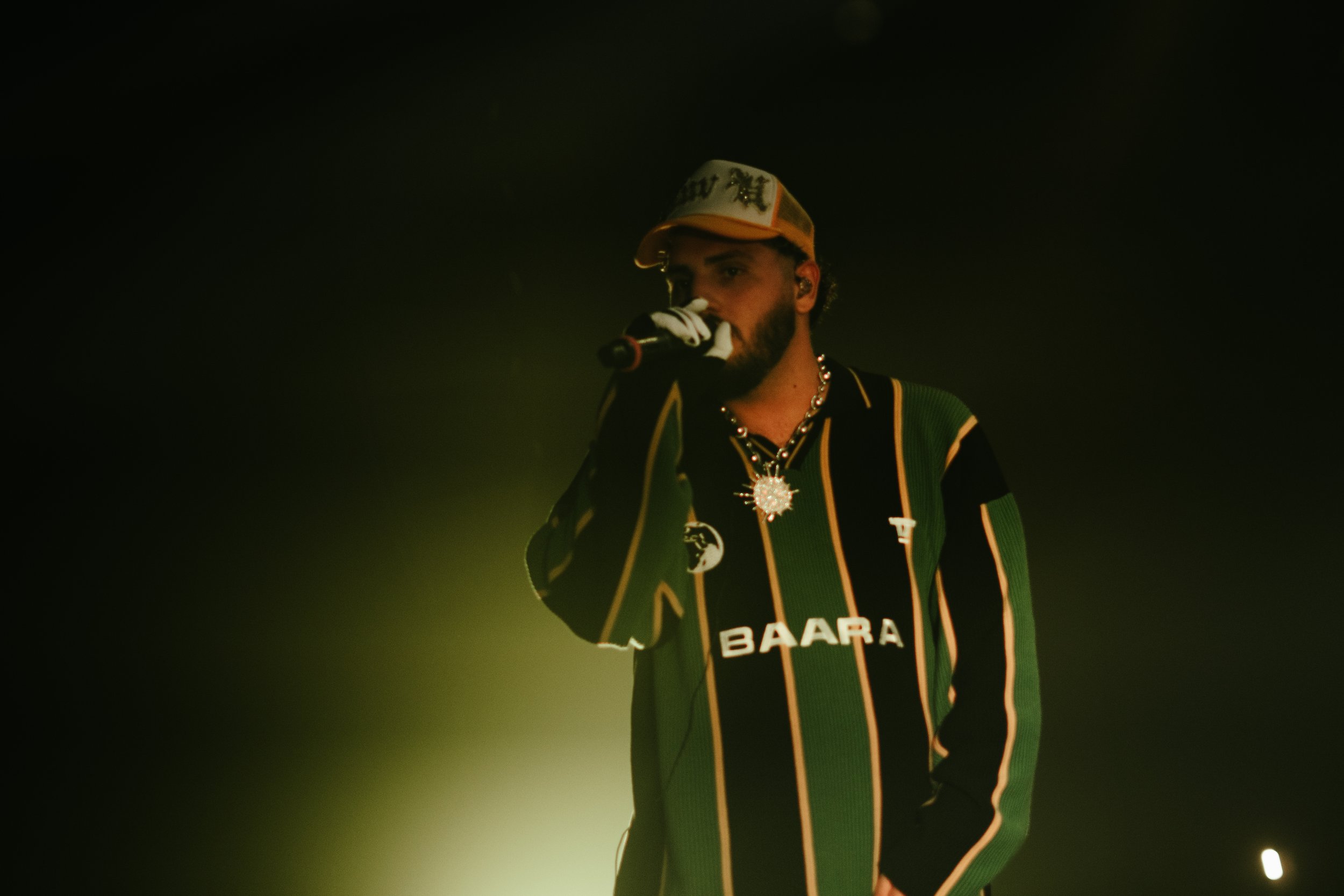 Un homme tenant un microphone, portant un bonnet, un collier et un sweat à rayures vertes, noires, jaunes et blanches, sur scène avec un éclairage faible.