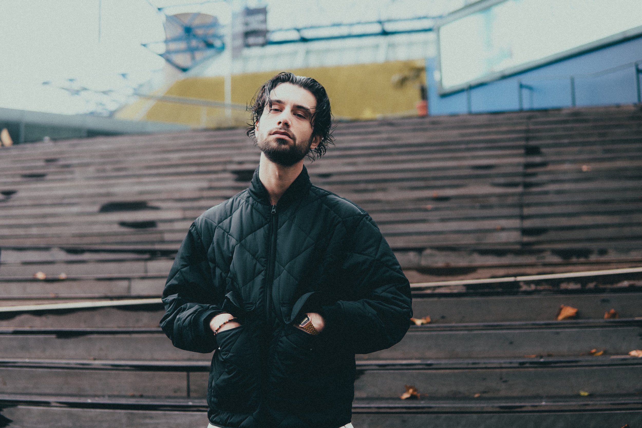 Un jeune homme avec des cheveux bruns longs et une barbe, portant une veste noire, se tenant devant des escaliers en bois à l'extérieur, avec un bâtiment coloré en arrière-plan.
