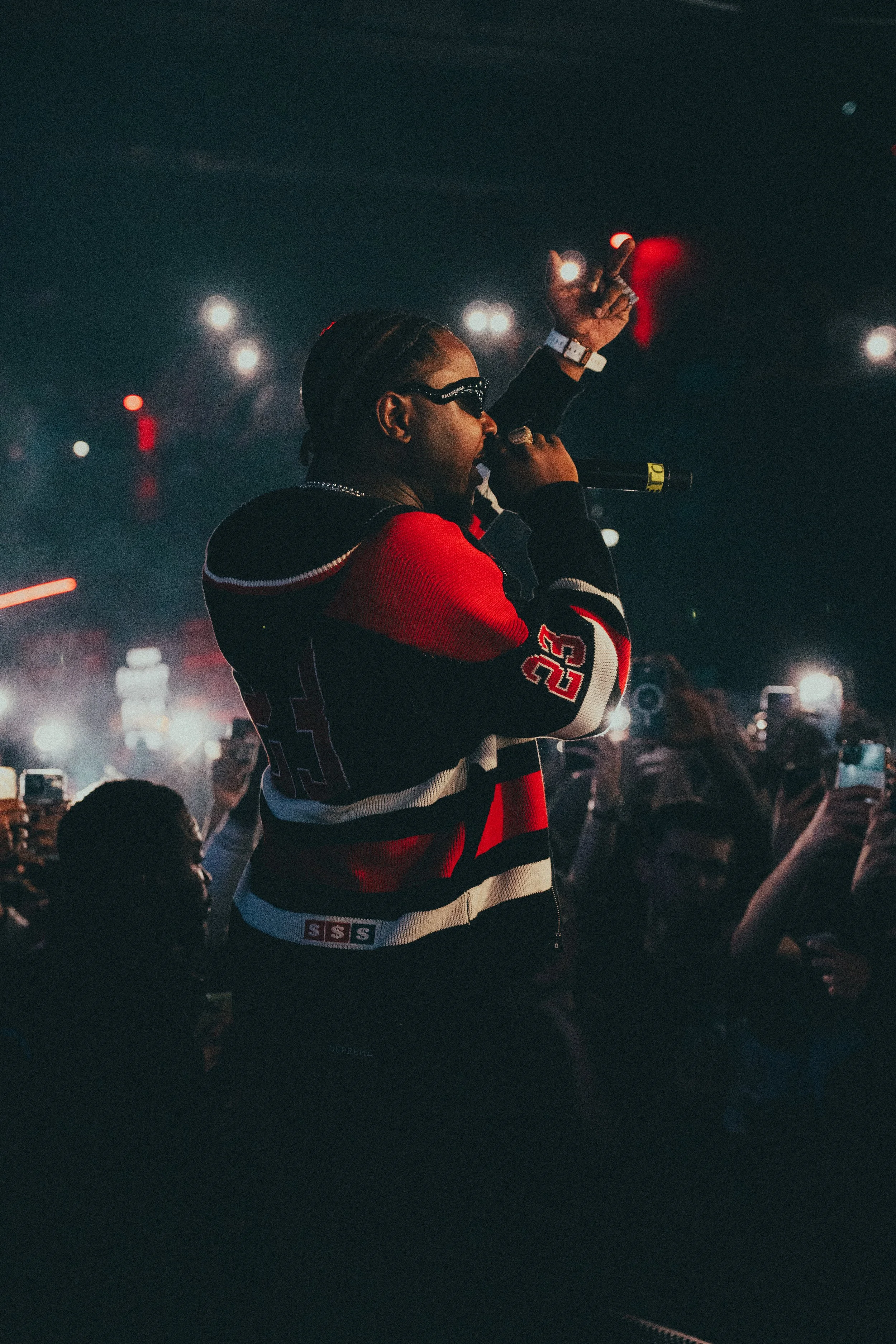 Un artiste en train de performer sur scène, tenant un micro et portant des lunettes de soleil, entouré d'une foule qui le filme avec leurs téléphones, lors d’un spectacle ou concert nocturne.