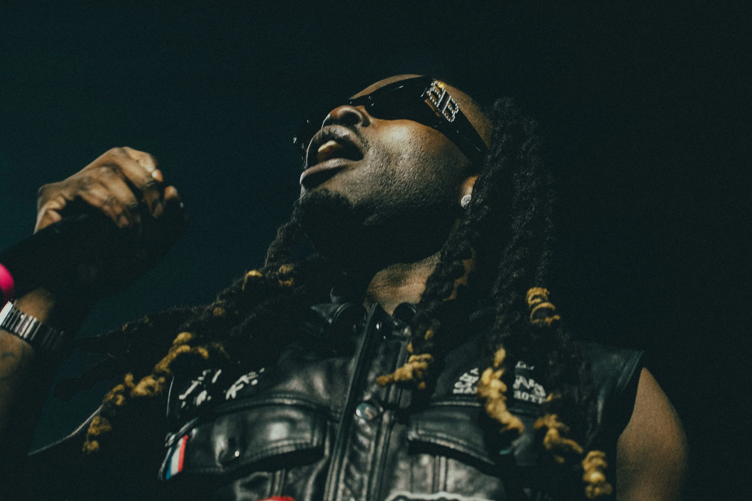 Un homme portant des lunettes de soleil noires, un blouson en cuir noir avec des détails en rouge, blanc, et bleu, et des dreadlocks, semble chanter ou parler avec passion dans un contexte sombre.