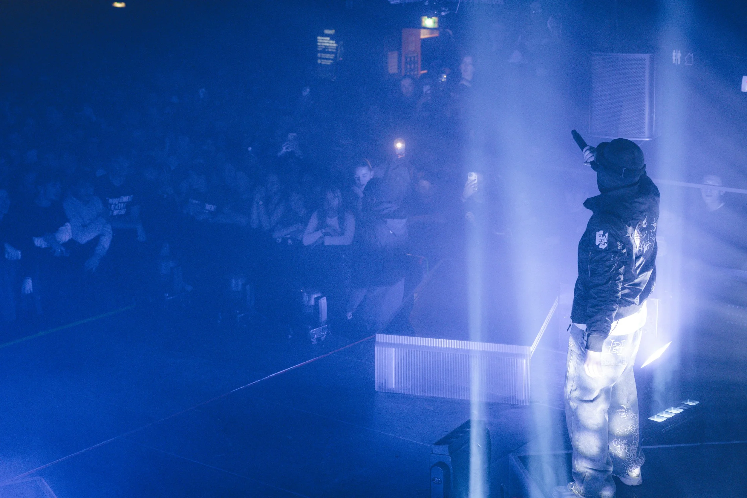 Un artiste de rue avec un chapeau de baseball fait un numéro de rapène devant un public dans un lieu sombre éclairé par des projecteurs bleus.