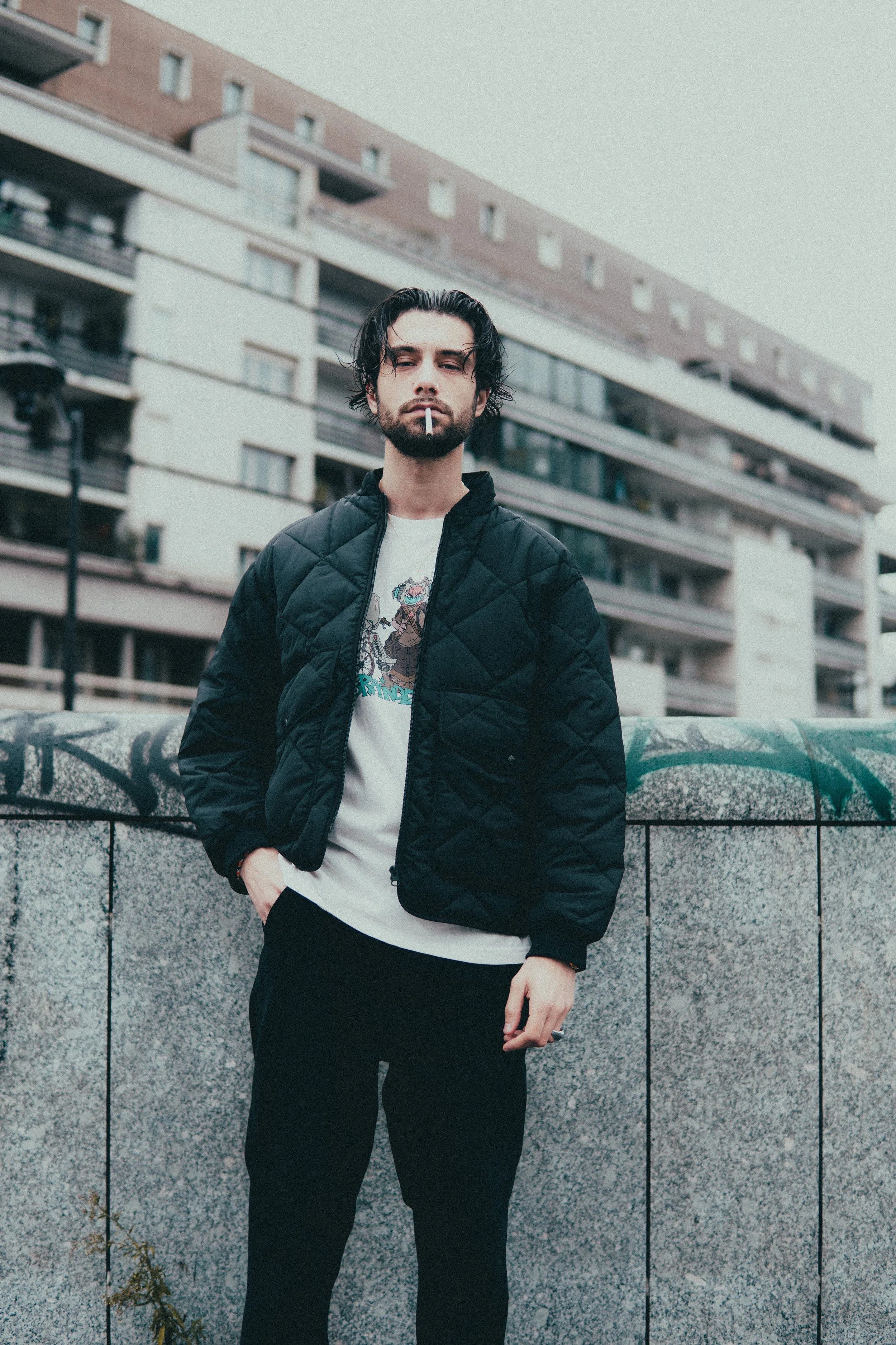 Un jeune homme avec des cheveux noirs et une barbe, portant une veste noire en nylon, une t-shirt blanc avec un dessin, tenant une cigarette dans la bouche, se tenant devant un mur en pierre avec du graffiti, avec des bâtiments résidentiels en arrièr